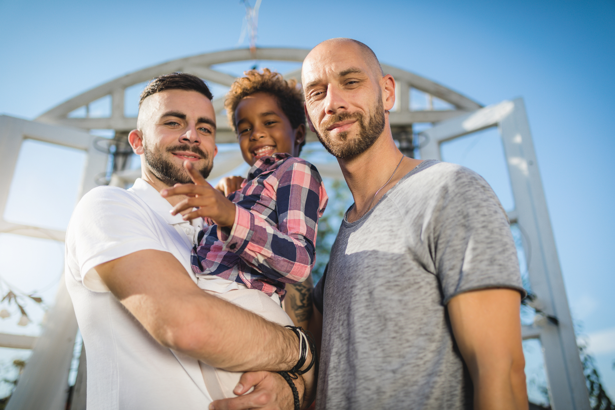 Two men with a child on shoulders smiling outdoors, expressing family closeness