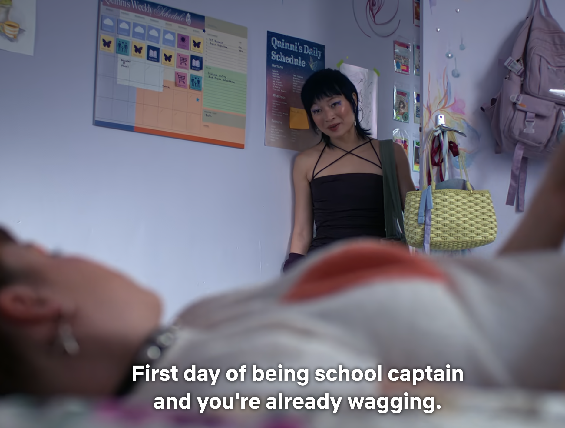 Person in school uniform lies on bed; another person with black hair, black top, and skirt stands nearby. Dialogue from TV show on screen
