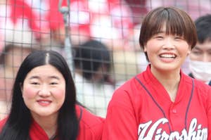 広島カープのユニフォームを着た2人の女性が野球場で笑顔で立っている。