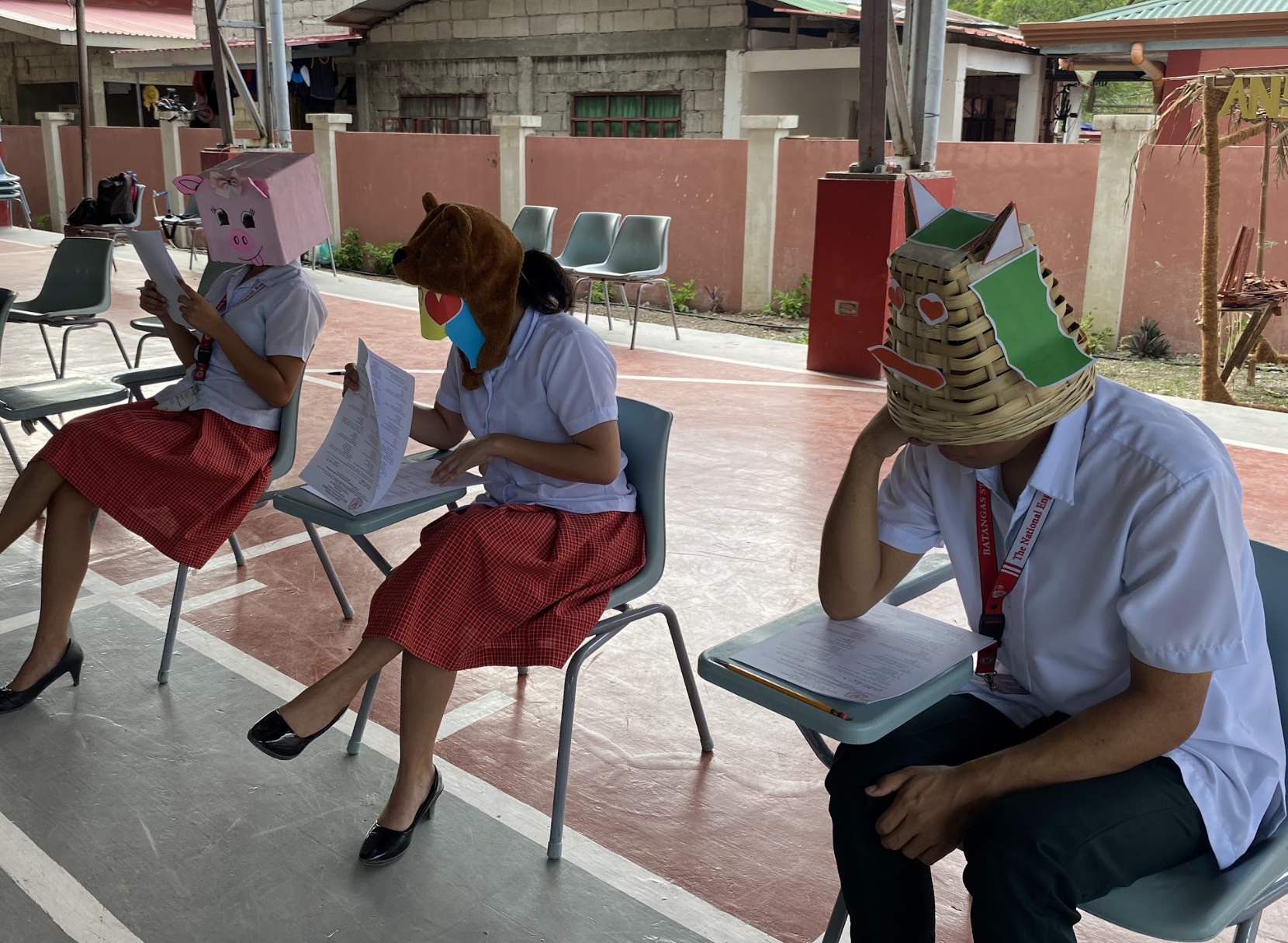 Three students wearing animal masks sit reading books outdoors, with one teacher supervising