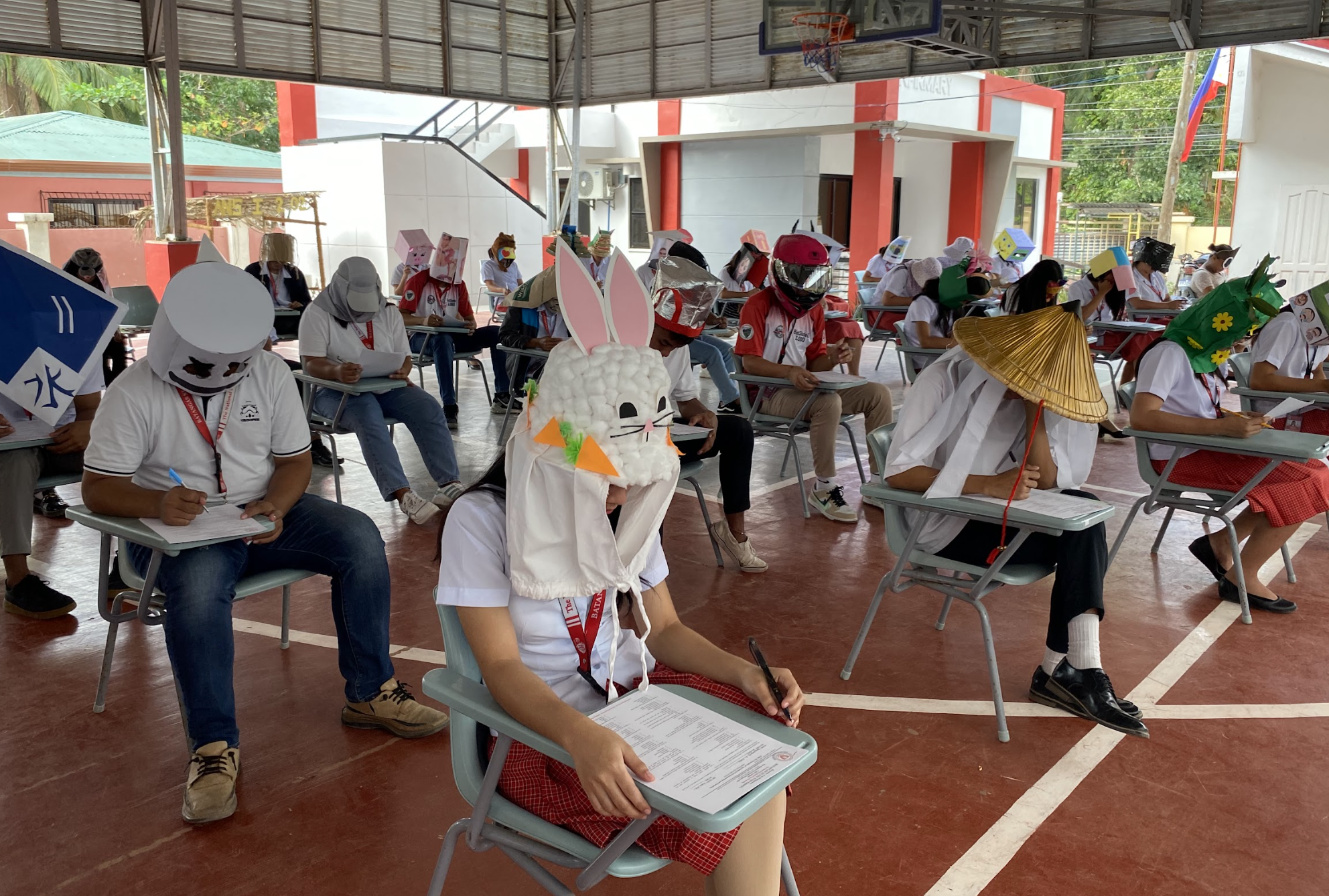 Students wearing various handmade masks seated at desks outdoors during an event