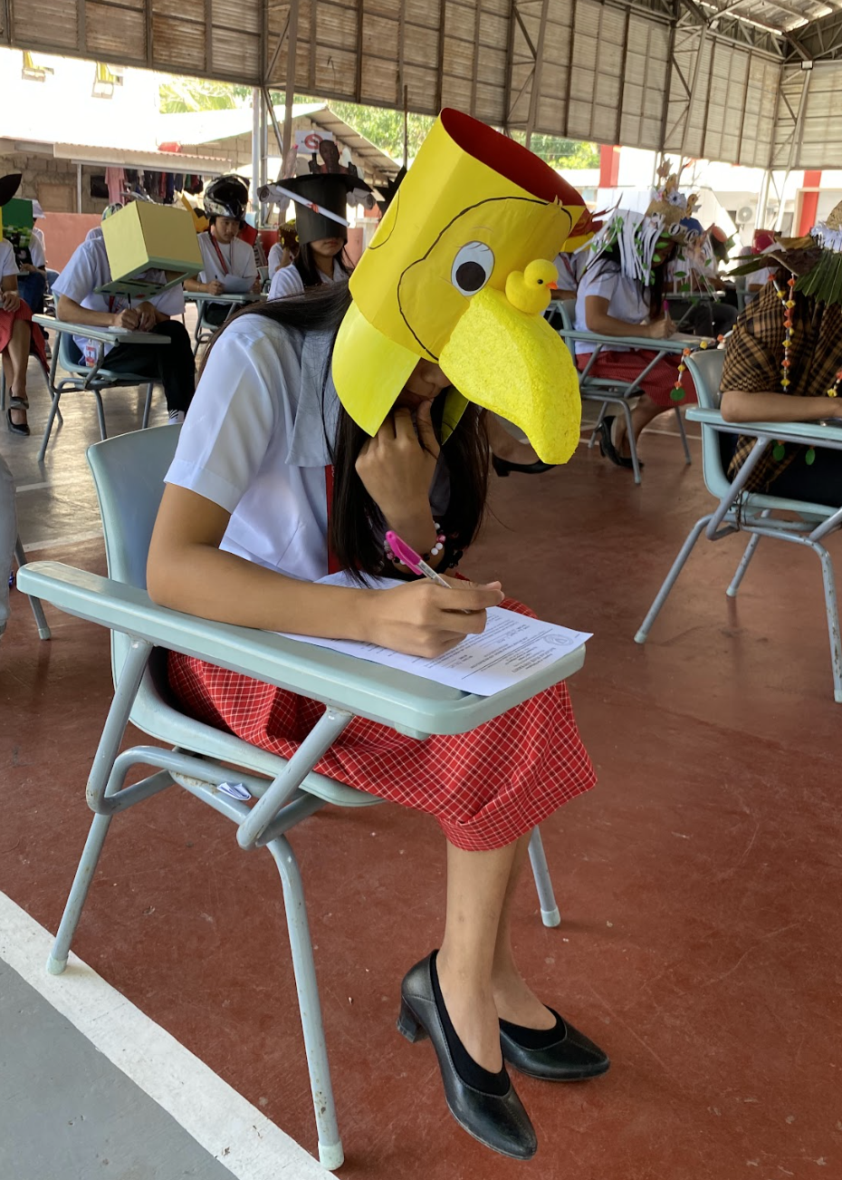 Student in a plaid skirt and black shoes wearing a large bird head mask while seated and writing