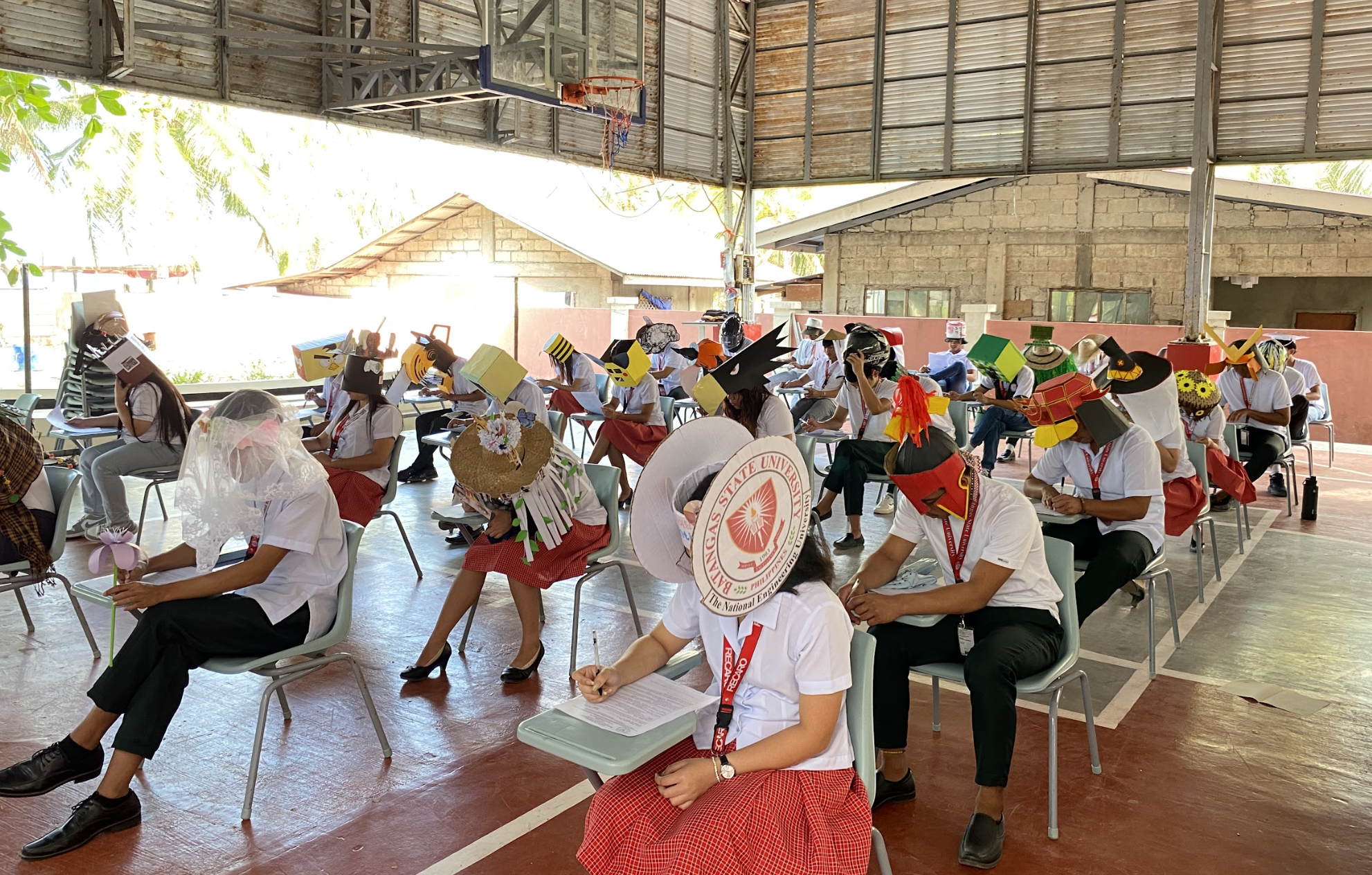 Students wearing face shields and various hats sit in a classroom setup with desks socially distanced