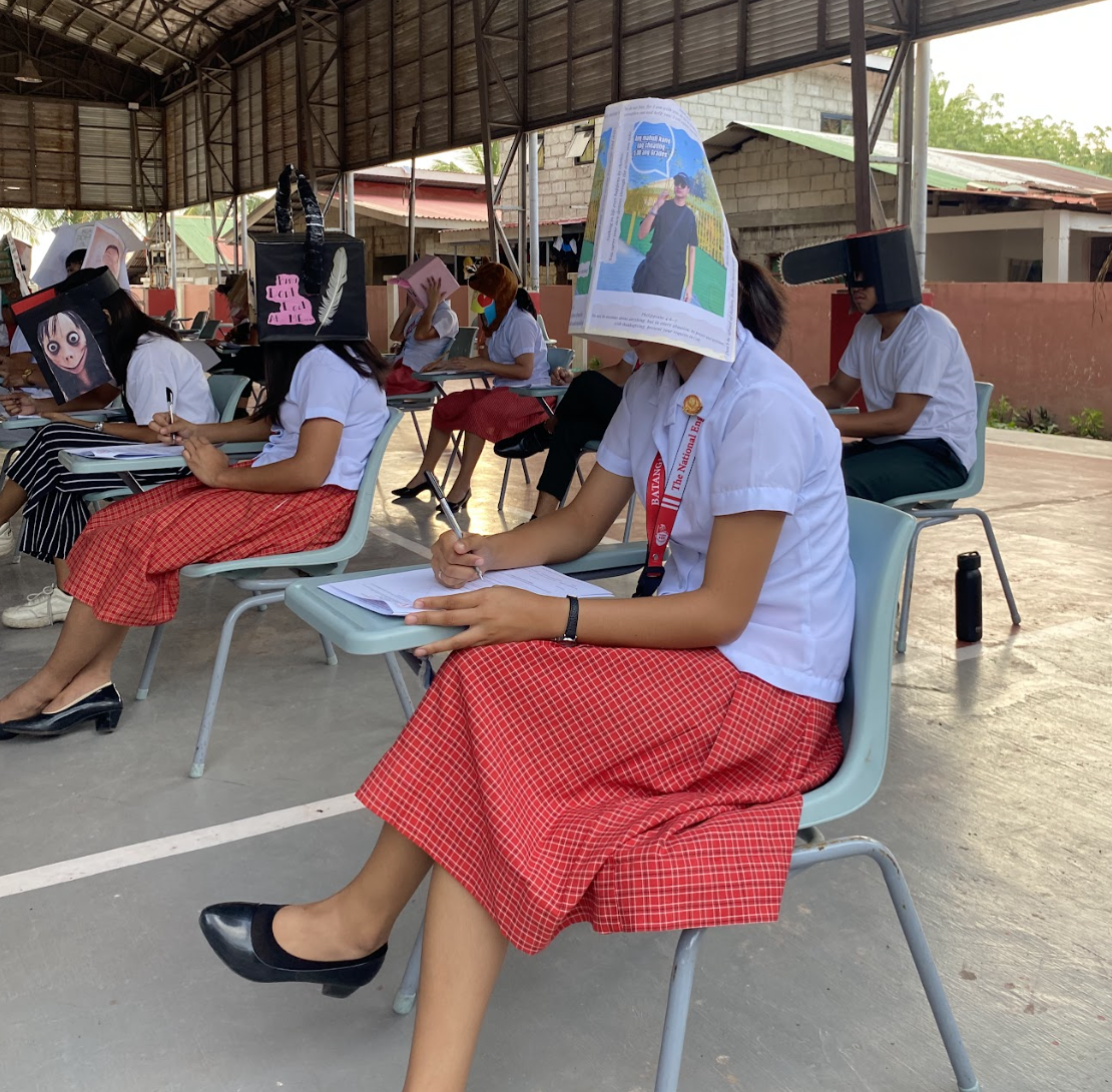 Students wearing paper box head coverings sit at desks taking an exam outdoors