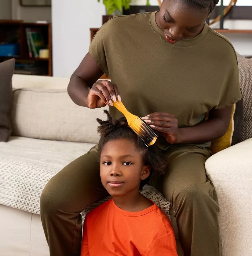Woman combing child's hair on couch, both dressed casually, in a home setting