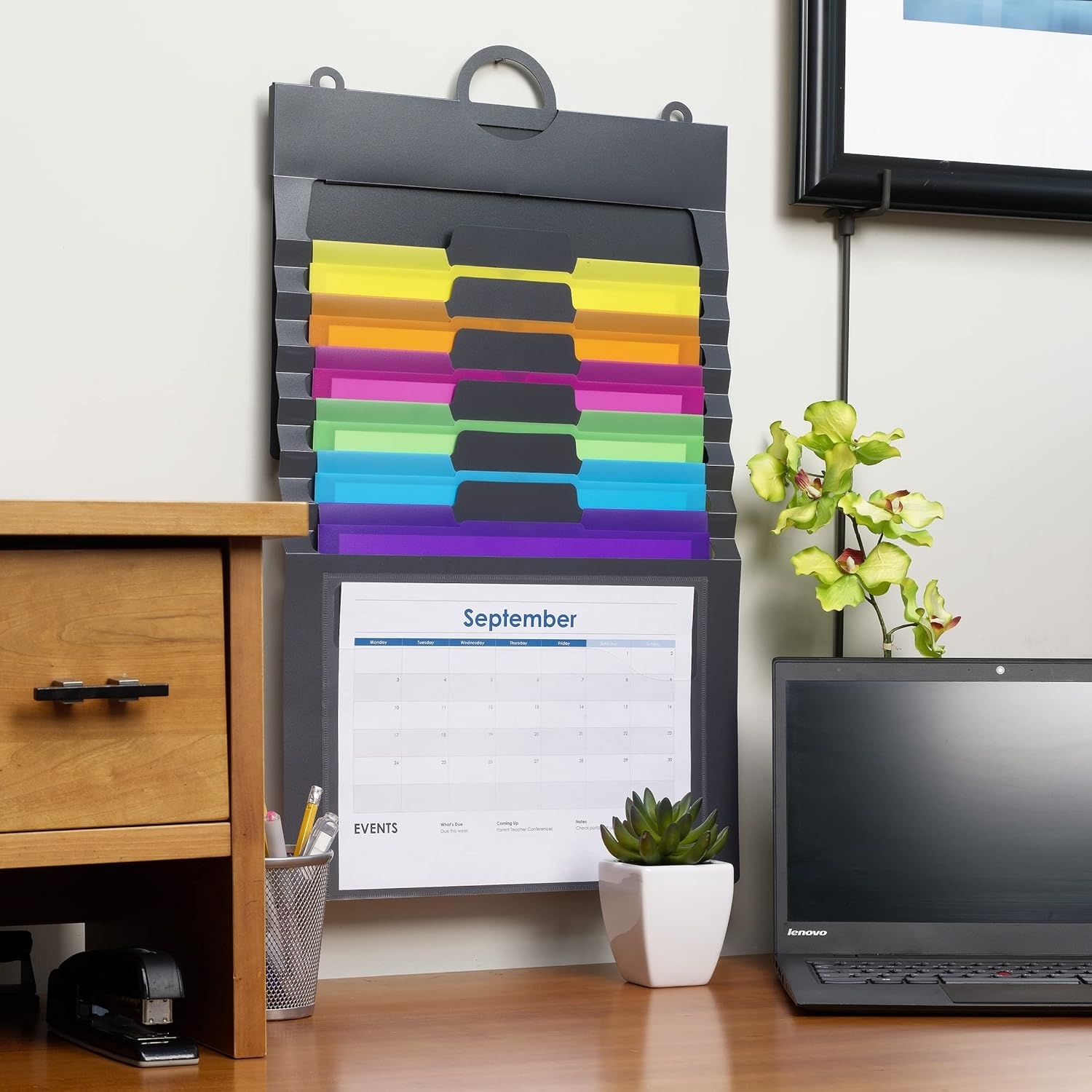 File organizer with colorful tabs attached to a wall above a desk with a calendar, laptop, and plants