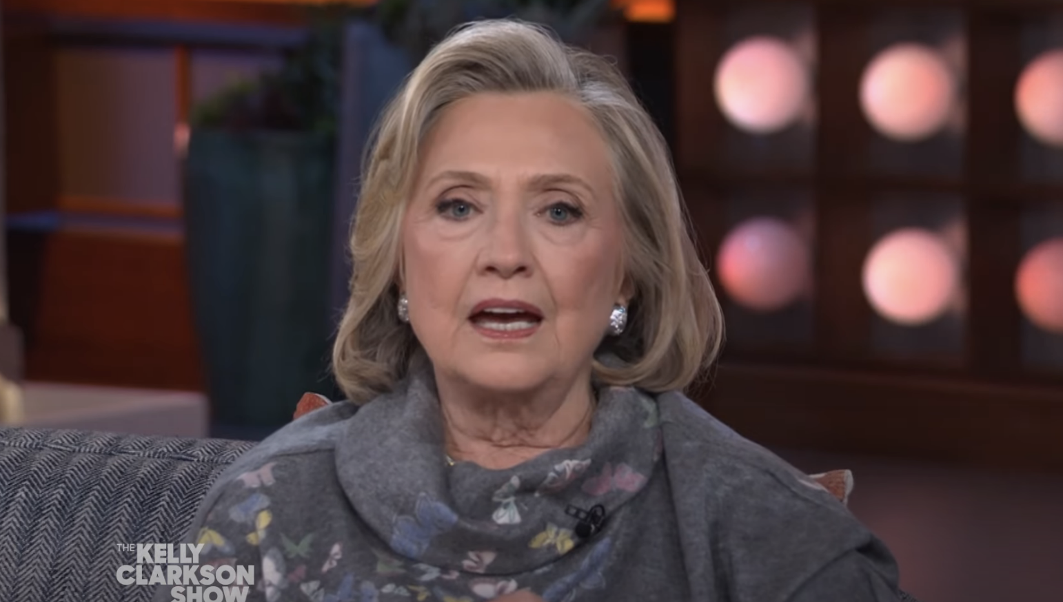 Hillary Clinton wearing a jacket and scarf, seated on the talk show