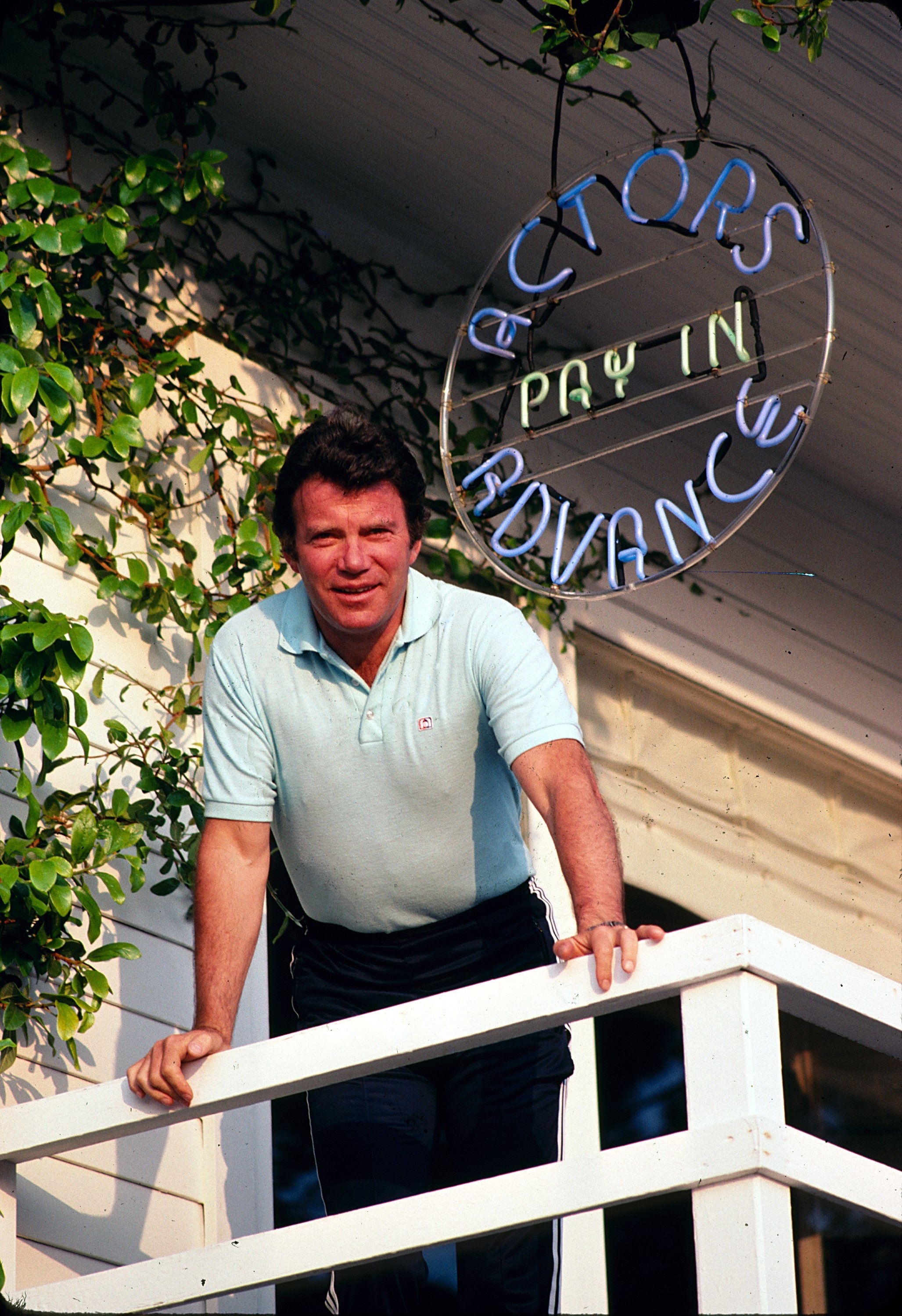 Man leaning on balcony rail below a neon sign that reads &quot;ACTORS PAY IN ADVANCE.&quot;