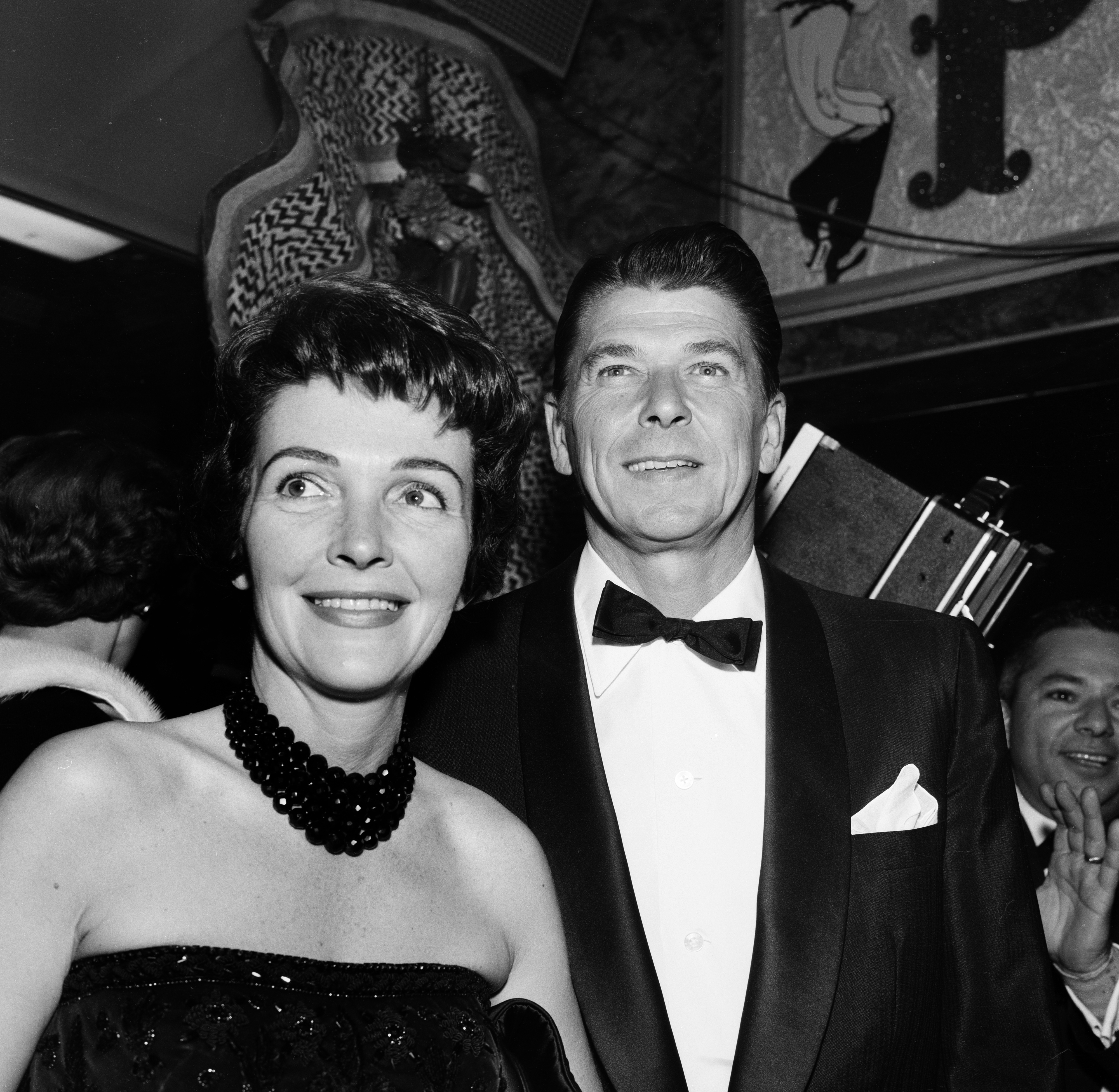Two individuals at an event, person on the right in a tuxedo with a bow tie, person on the left in a strapless gown with a necklace