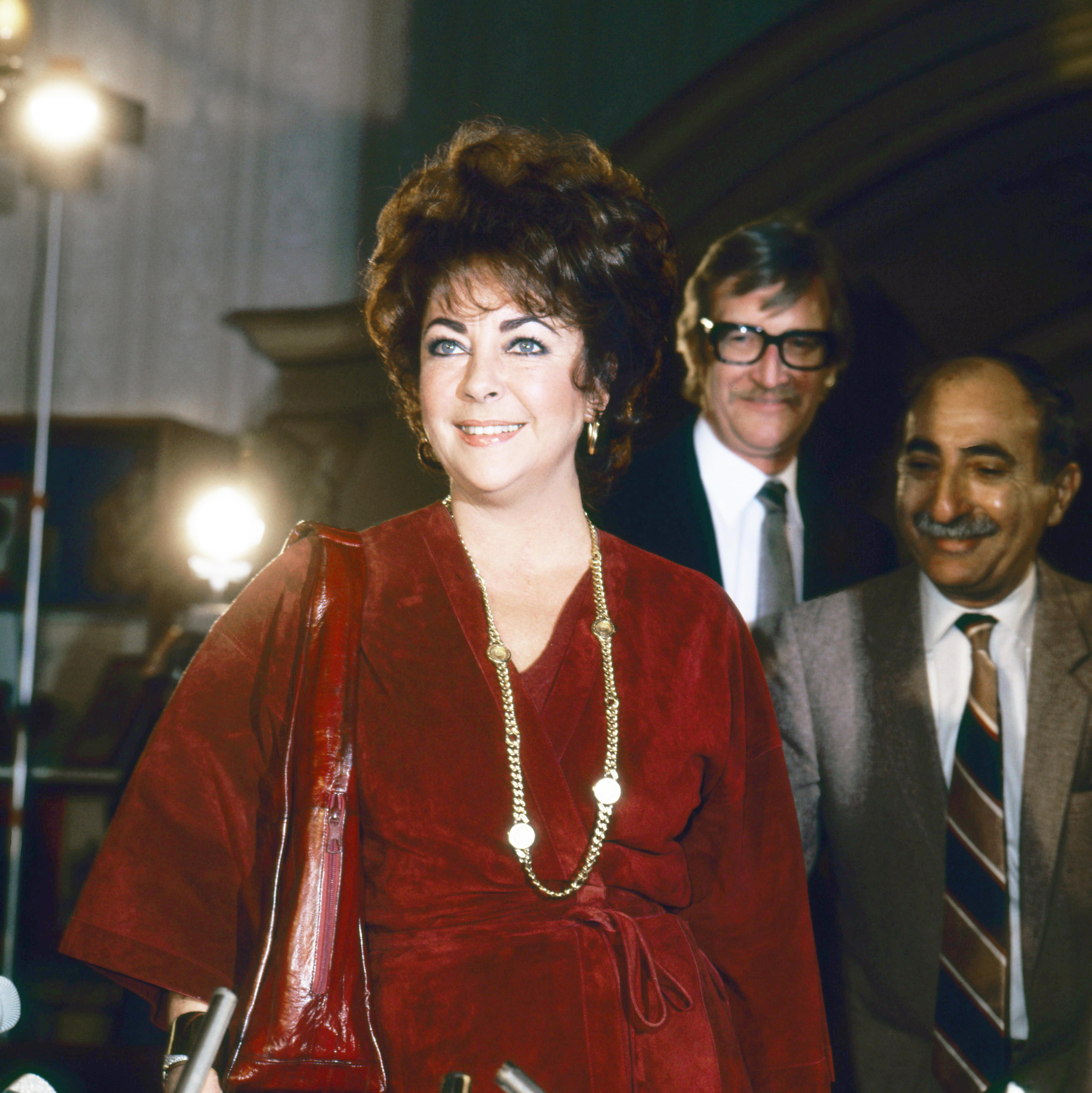 Woman in a fur coat and necklace, with two men in suits behind her