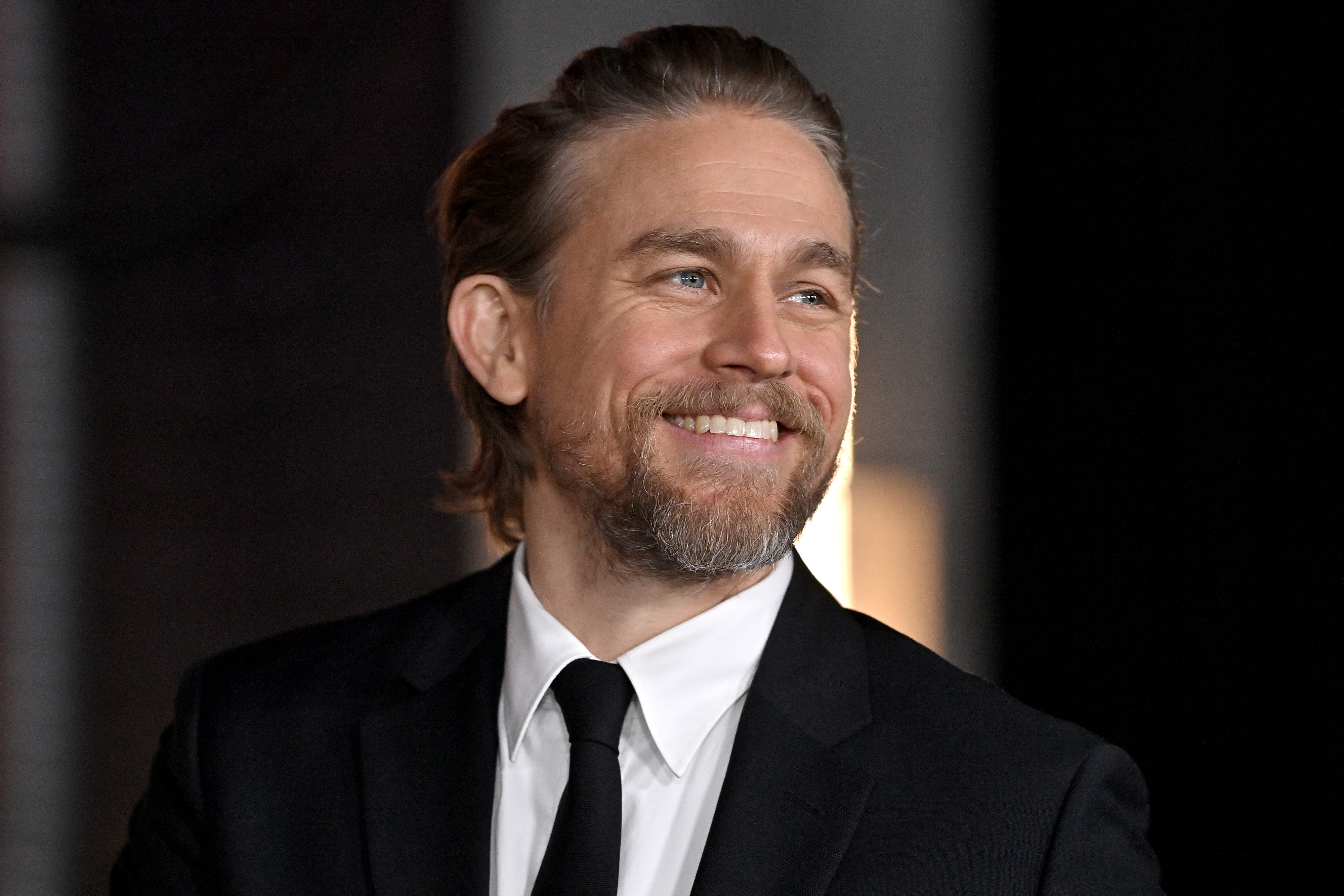Charlie Hunnam smiling while wearing a dark suit and tie