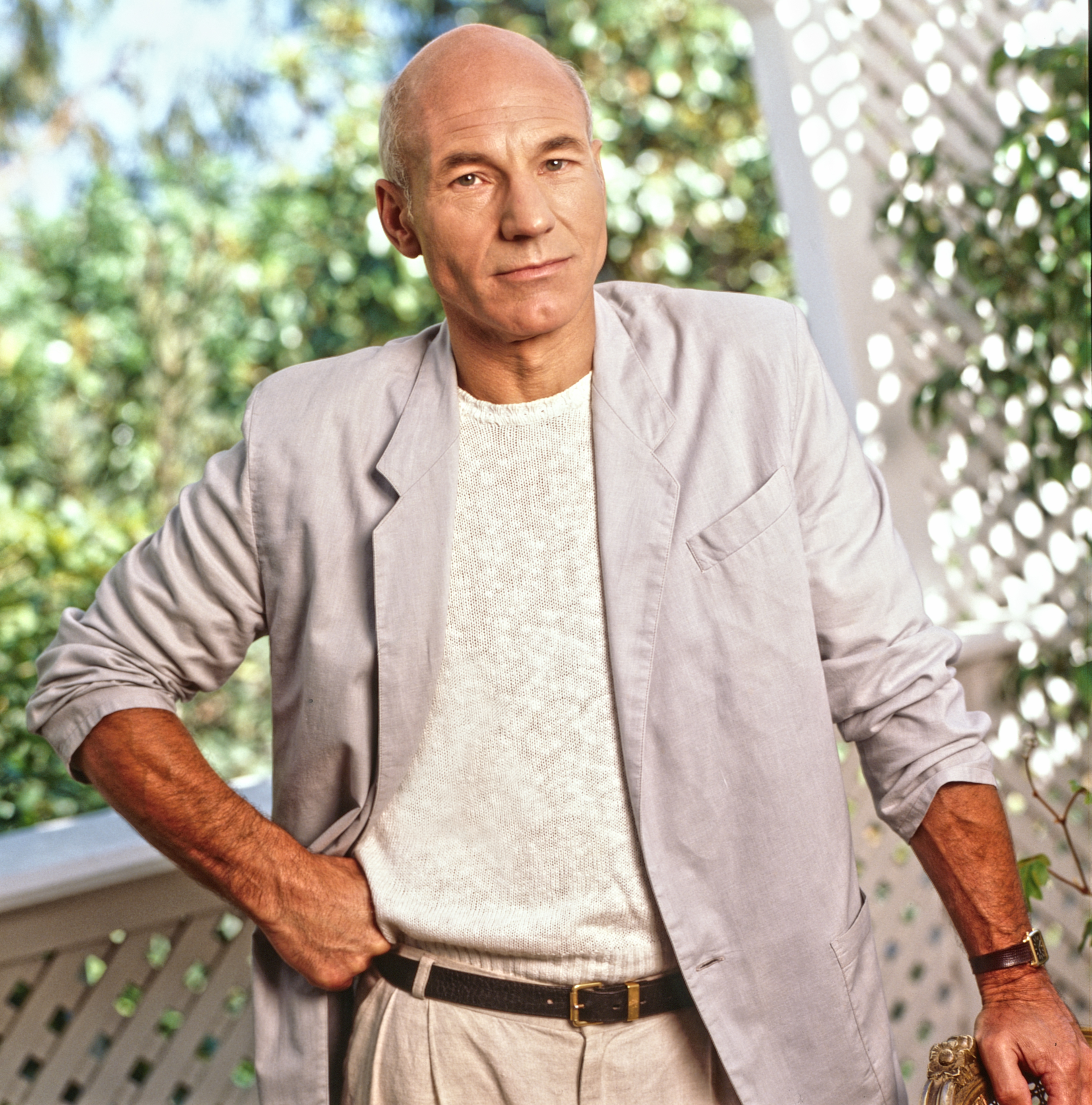 Patrick Stewart stands with hands on hips, wearing a light jacket and sweater