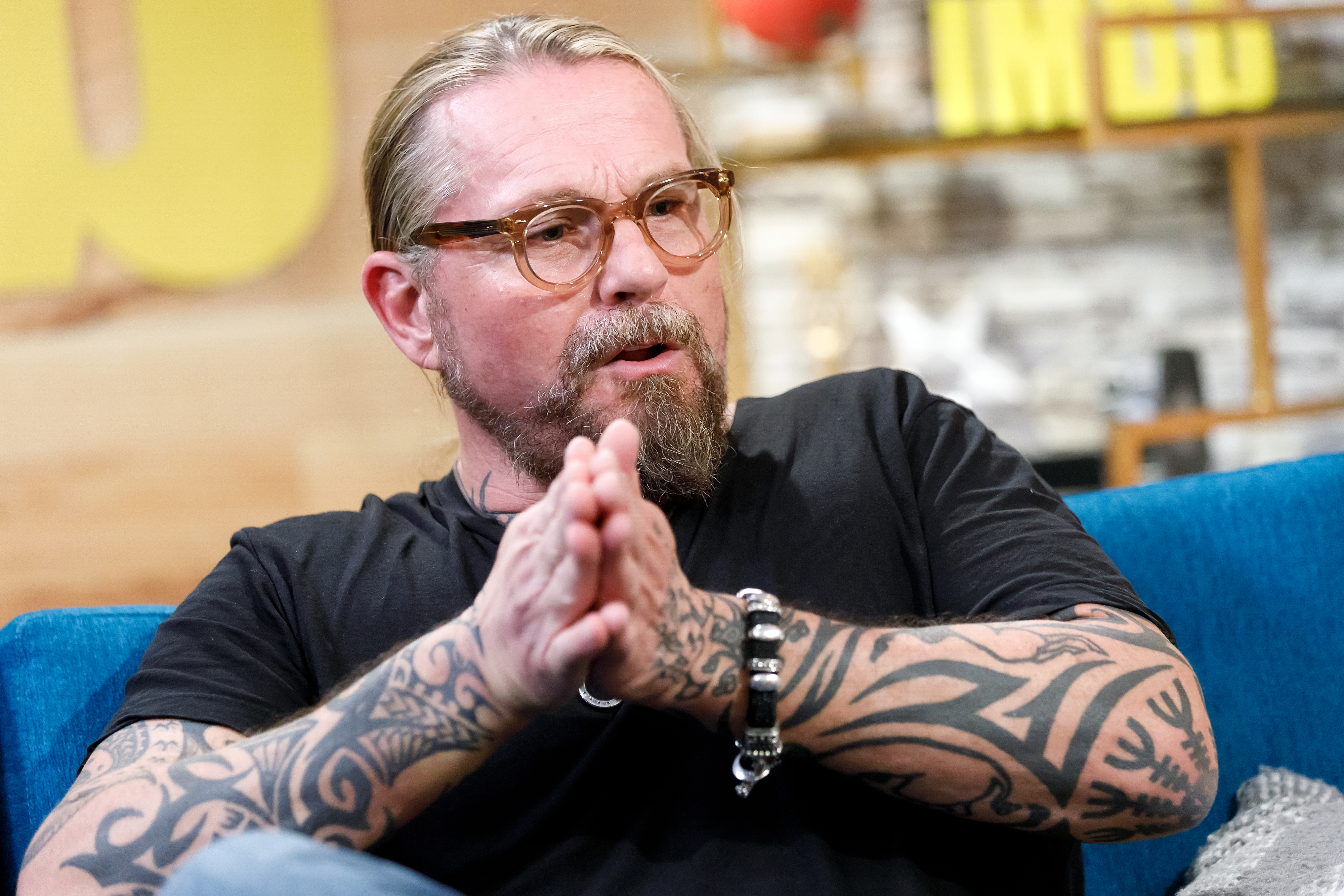 Kurt Sutter with beard and tattoos wearing glasses is seated with his hands clasped