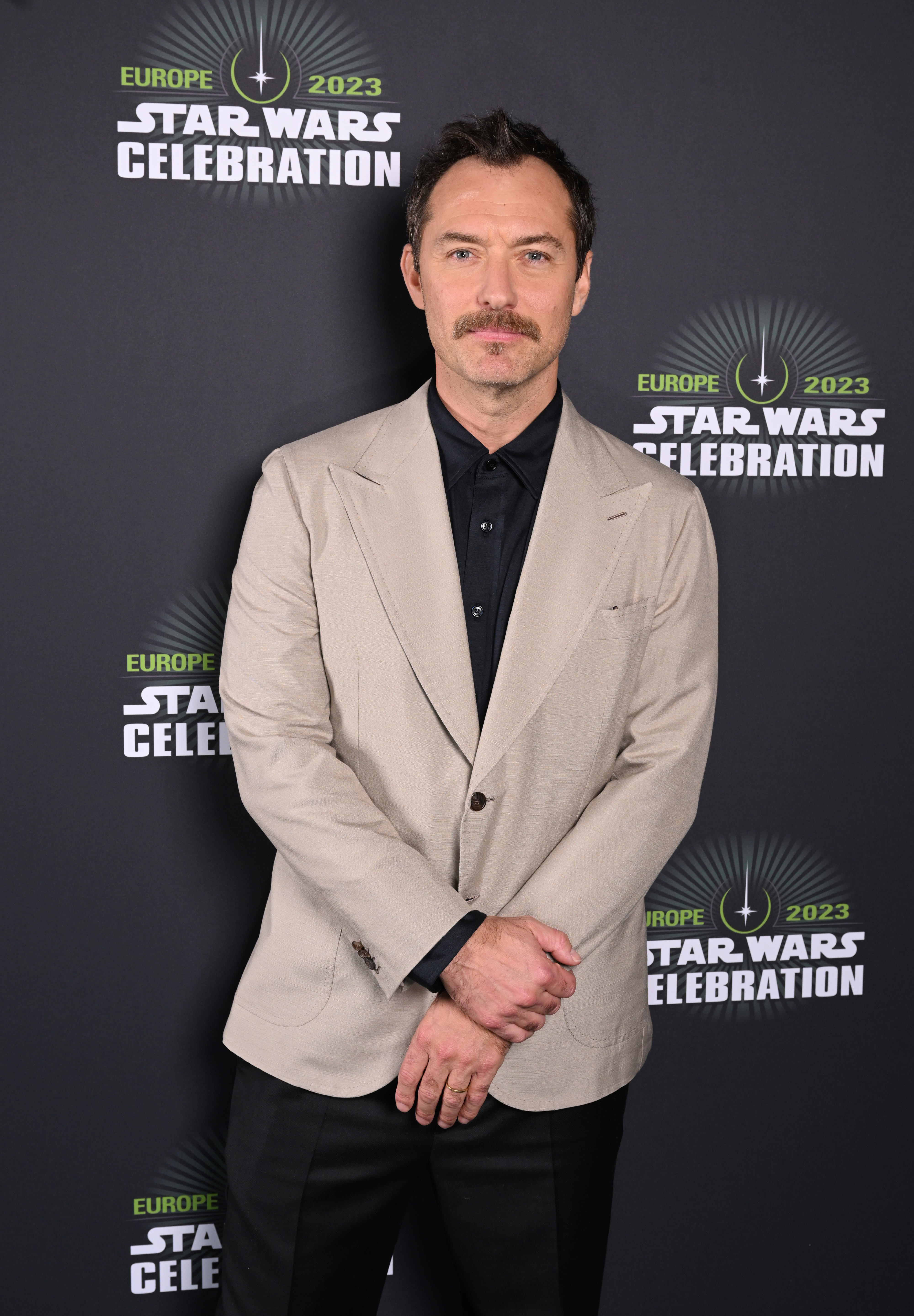 Ewan McGregor stands with hands clasped, wearing a beige blazer over a black shirt at the Star Wars Celebration event