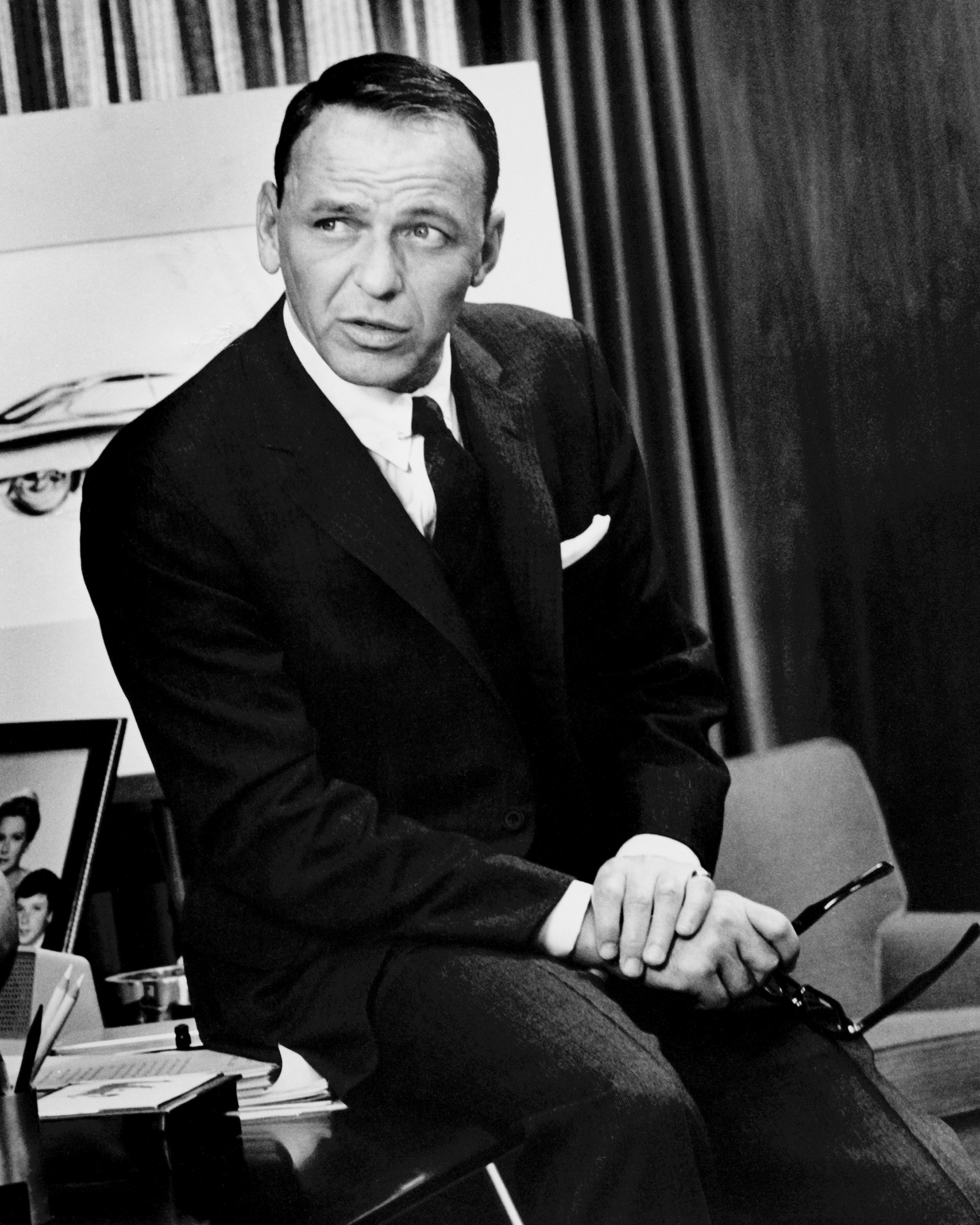 Man in suit seated with crossed legs, leaning forward, against backdrop with framed pictures