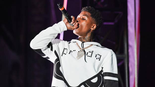 NBA YoungBoy performing on stage with a microphone, wearing a white graphic sweatshirt and chain necklace