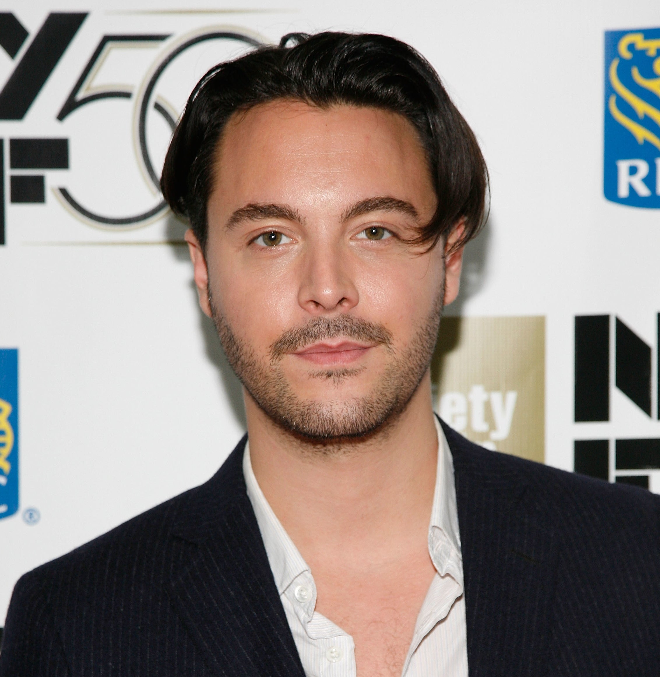 Jack Huston in dark blazer over shirt at an event with sponsor logos in background