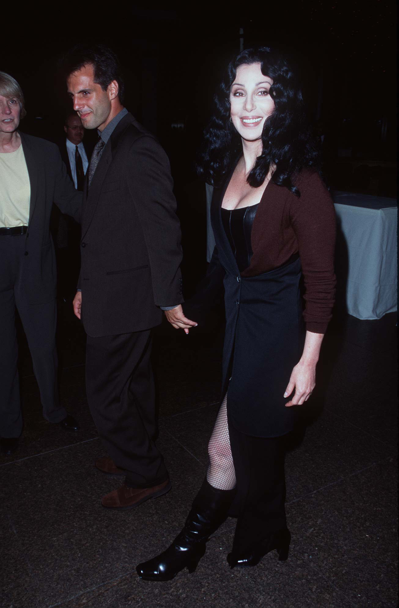 Cher in a black dress and boots, smiling as she walks with a male companion at an event
