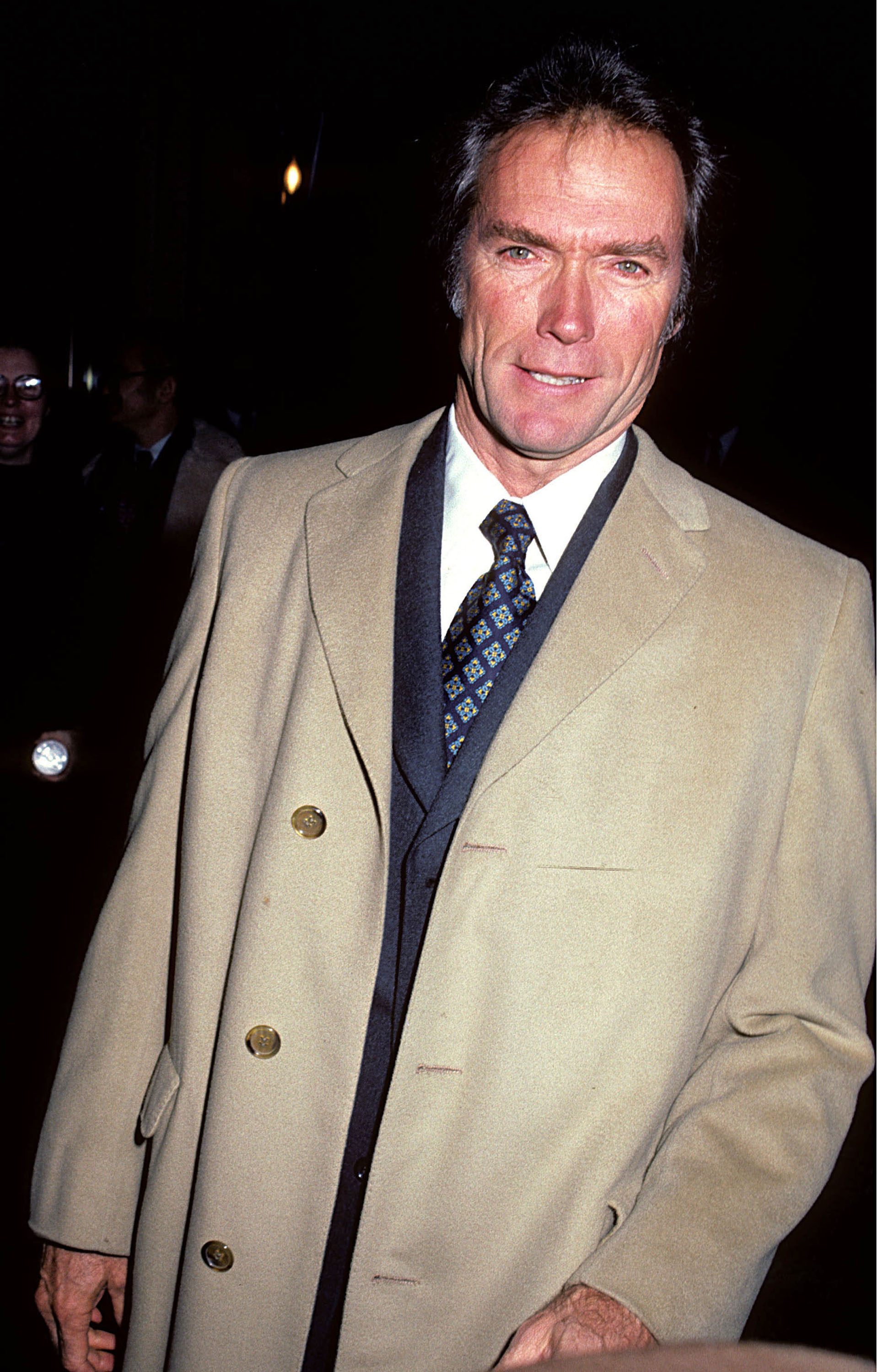 A person in a beige overcoat and patterned tie smiles at a camera