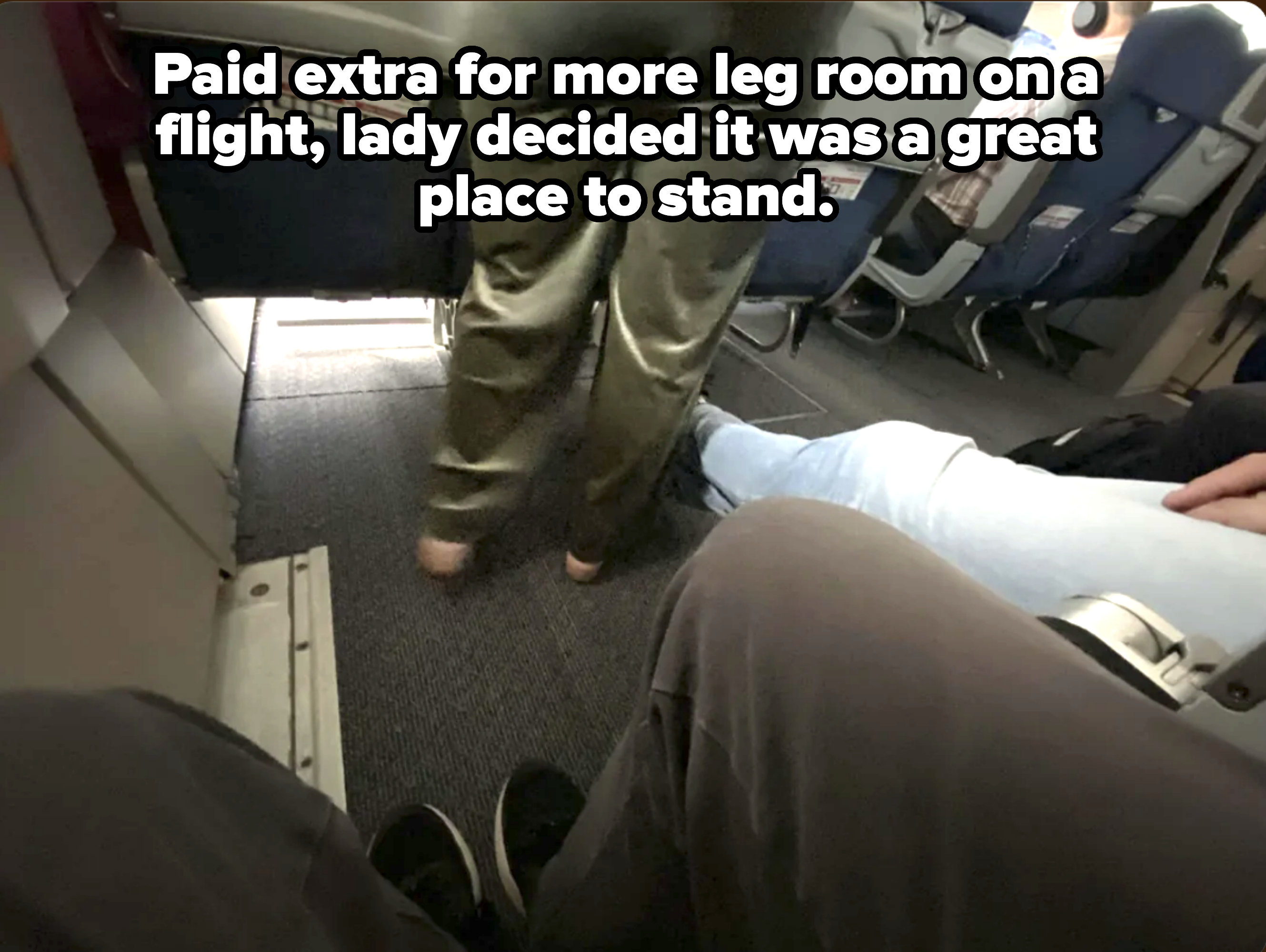 Passenger standing in airplane aisle next to seated passengers' legs, perspective from sitting down