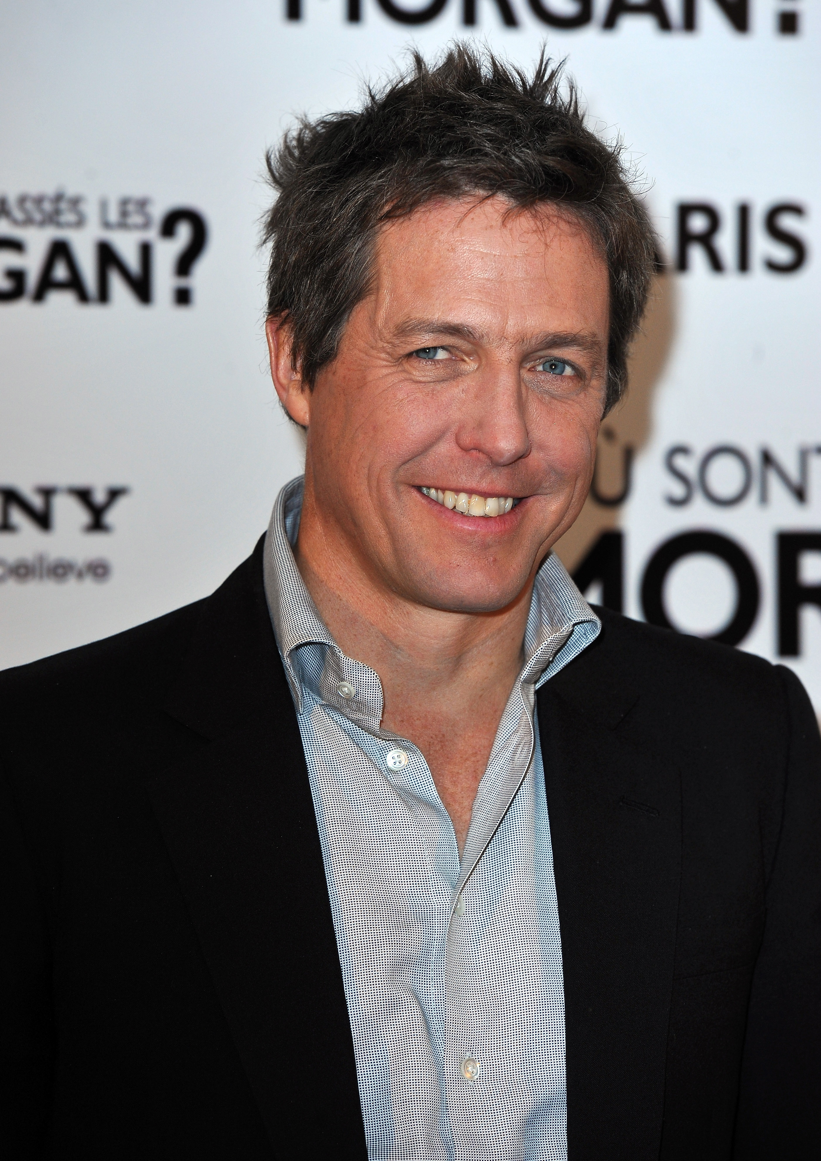 Hugh Grant wearing a dark jacket and light shirt, smiling at an event