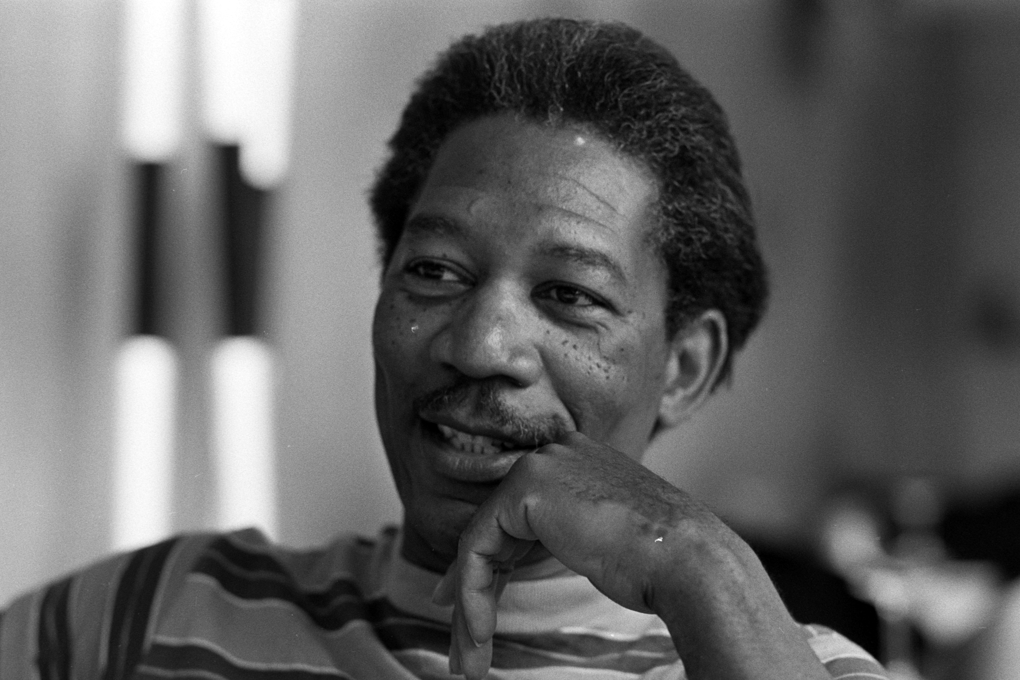 Morgan Freeman in a striped shirt, resting chin on hand with a thoughtful expression