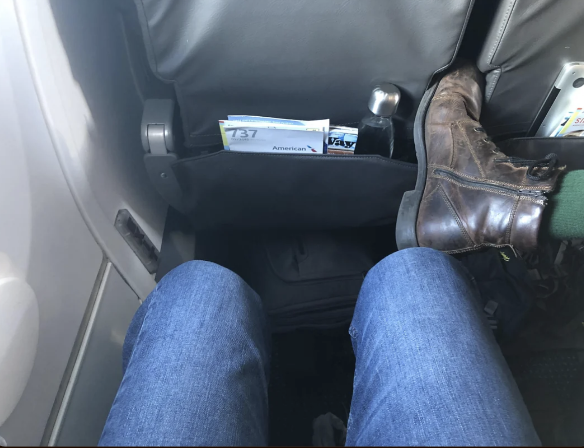 Person&#x27;s legs with jeans in an airplane seat, safety card in seatback pocket, and a brown boot on the right