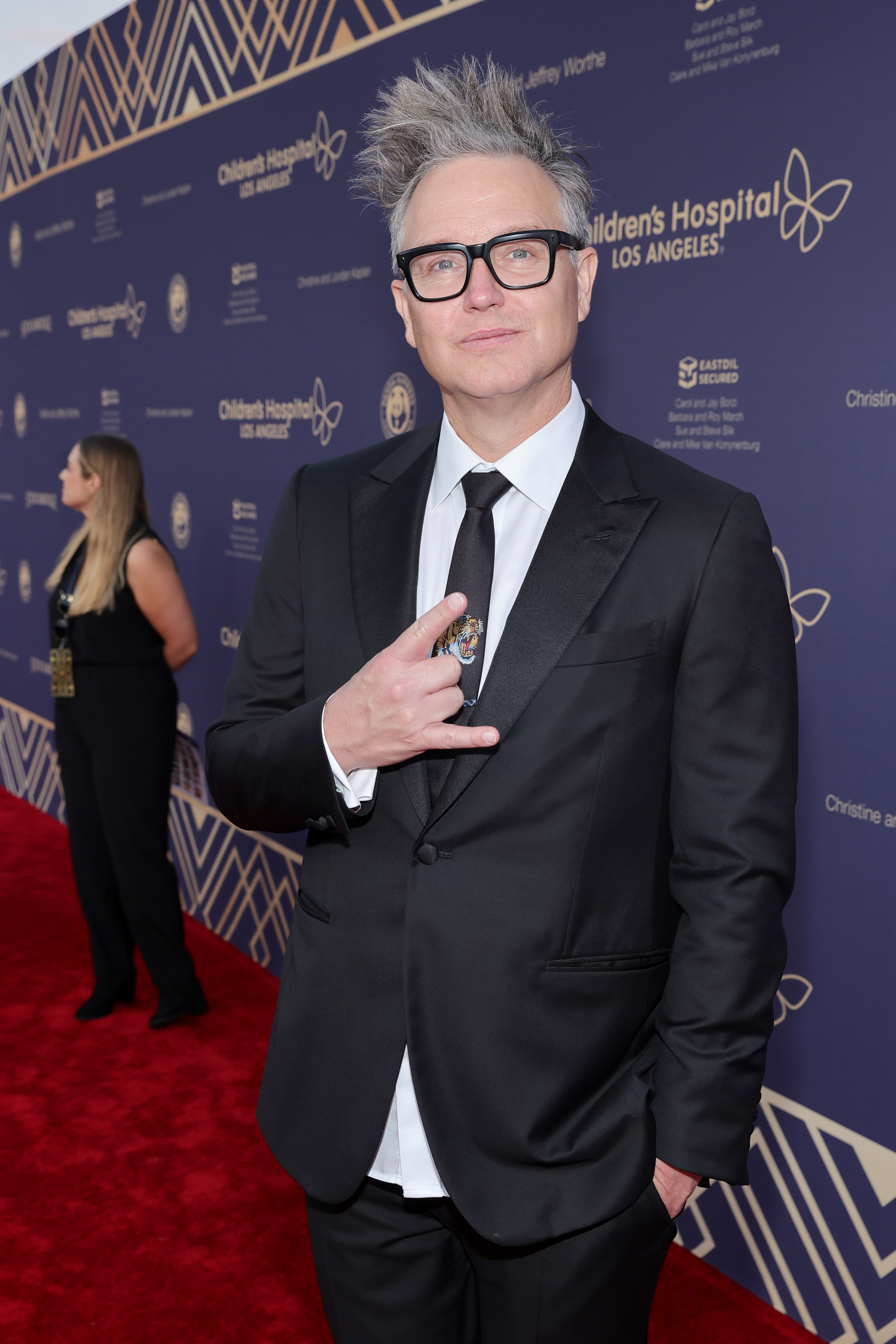 Person in a black suit and glasses posing with a peace sign on the red carpet
