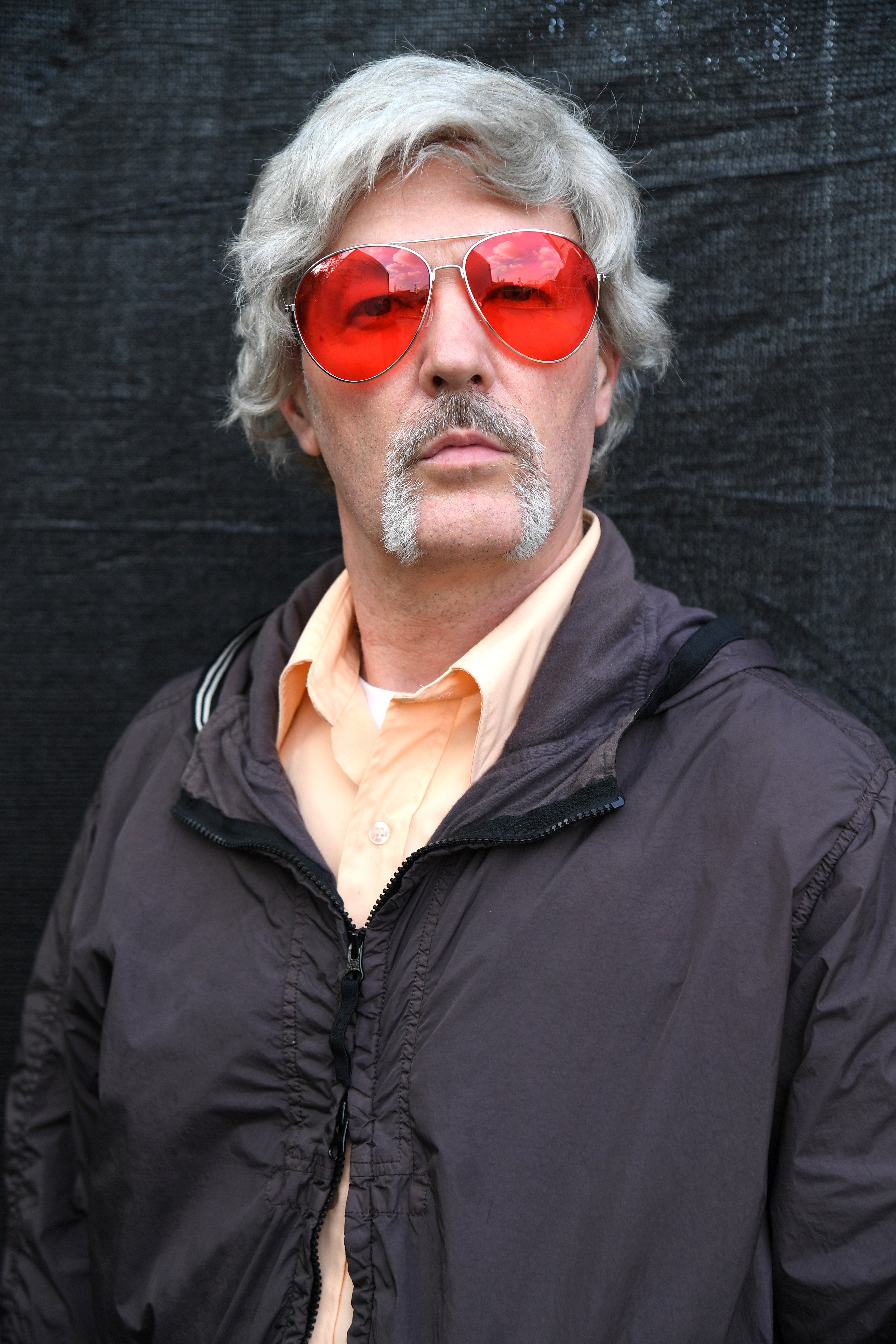 Man with heart-shaped sunglasses and a mustache, wearing a jacket over a collared shirt