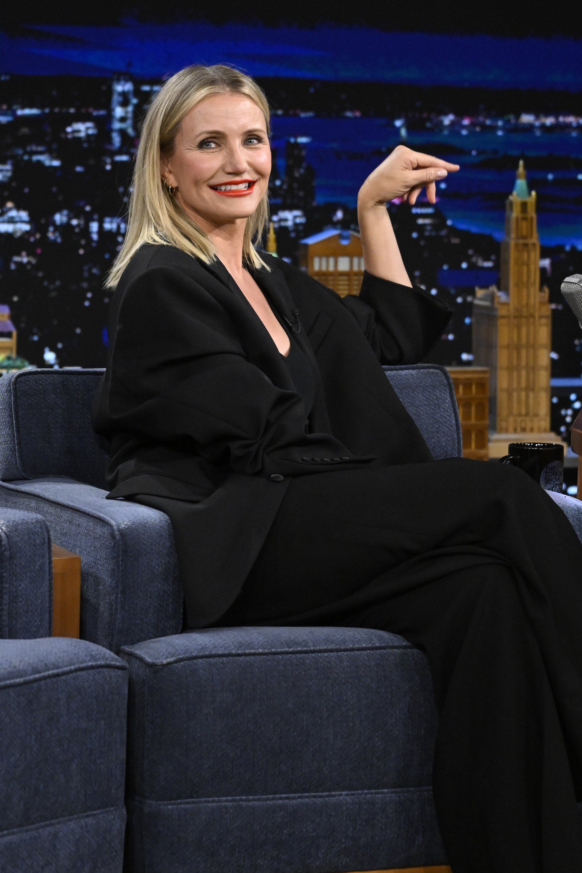 Woman in a black suit sits on a chair on a talk show set, smiling and pointing to her side