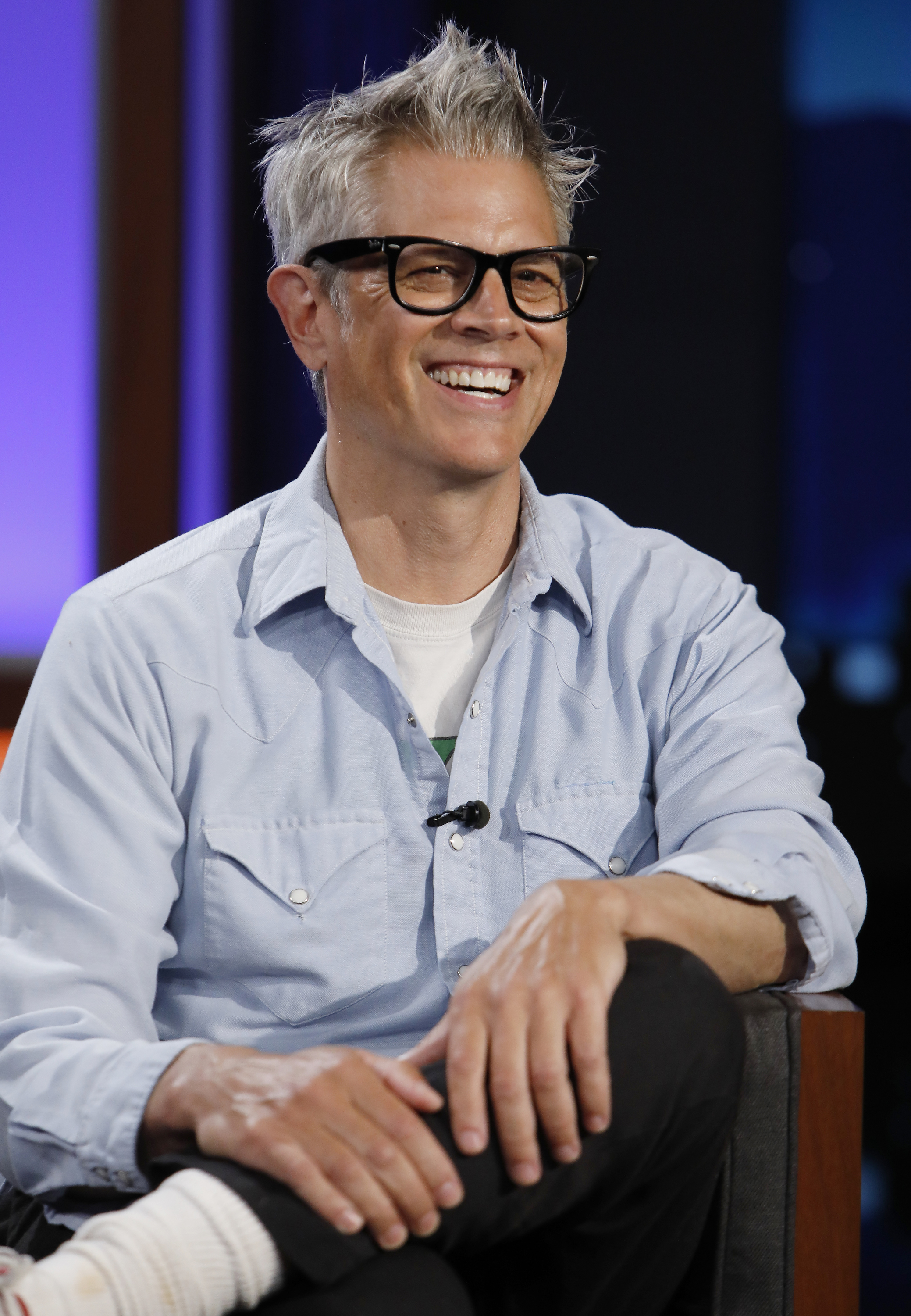 Person with short hair and glasses, smiling, seated in casual attire with layered shirts