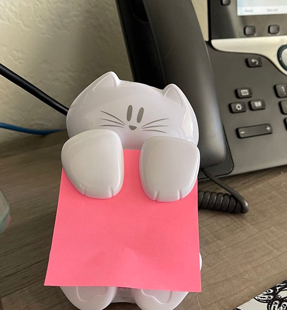 A cat-shaped note holder with a pink sticky note on a desk near a phone