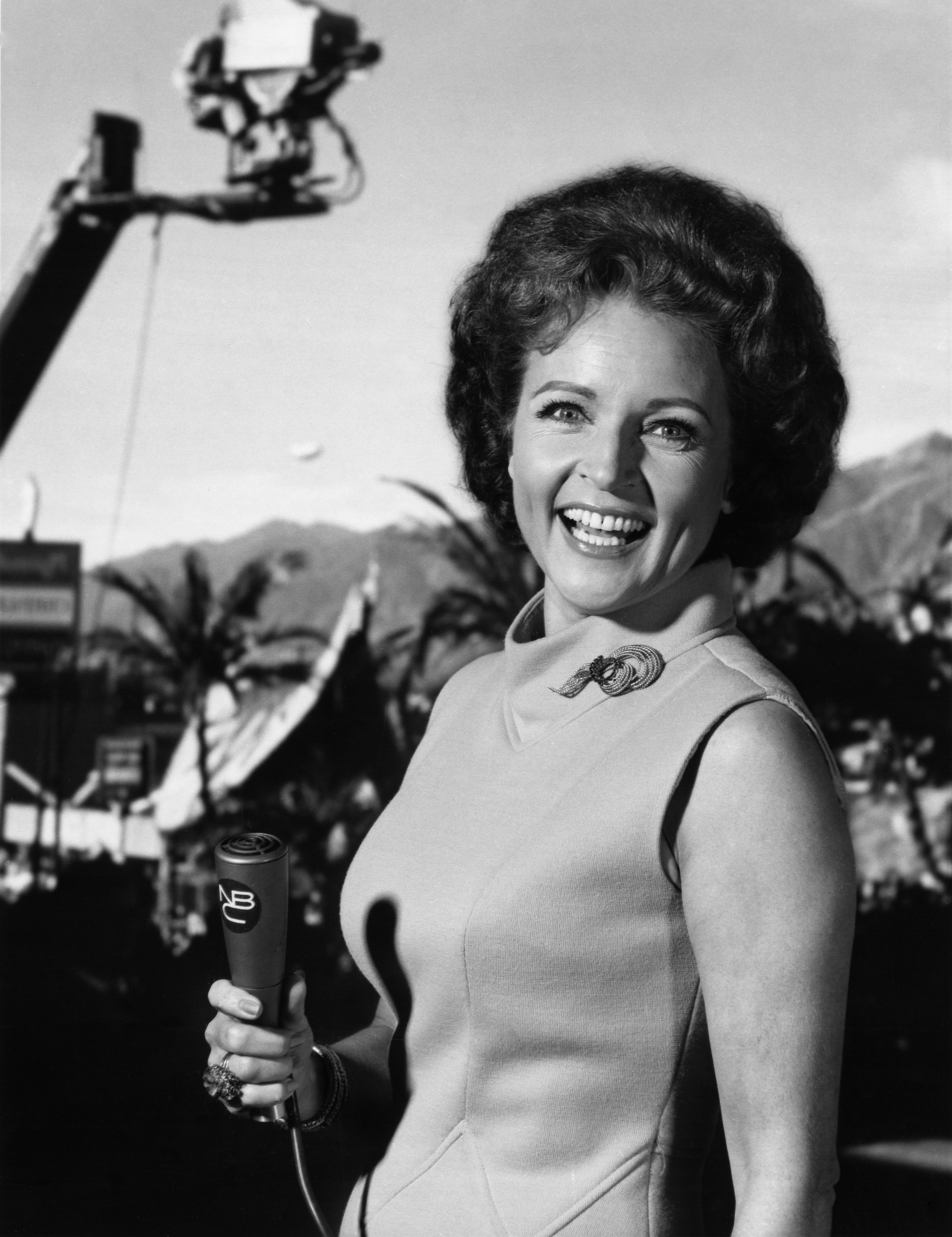 Woman in a sleeveless dress with a rose brooch, holding a microphone, smiling with a camera crane in the background