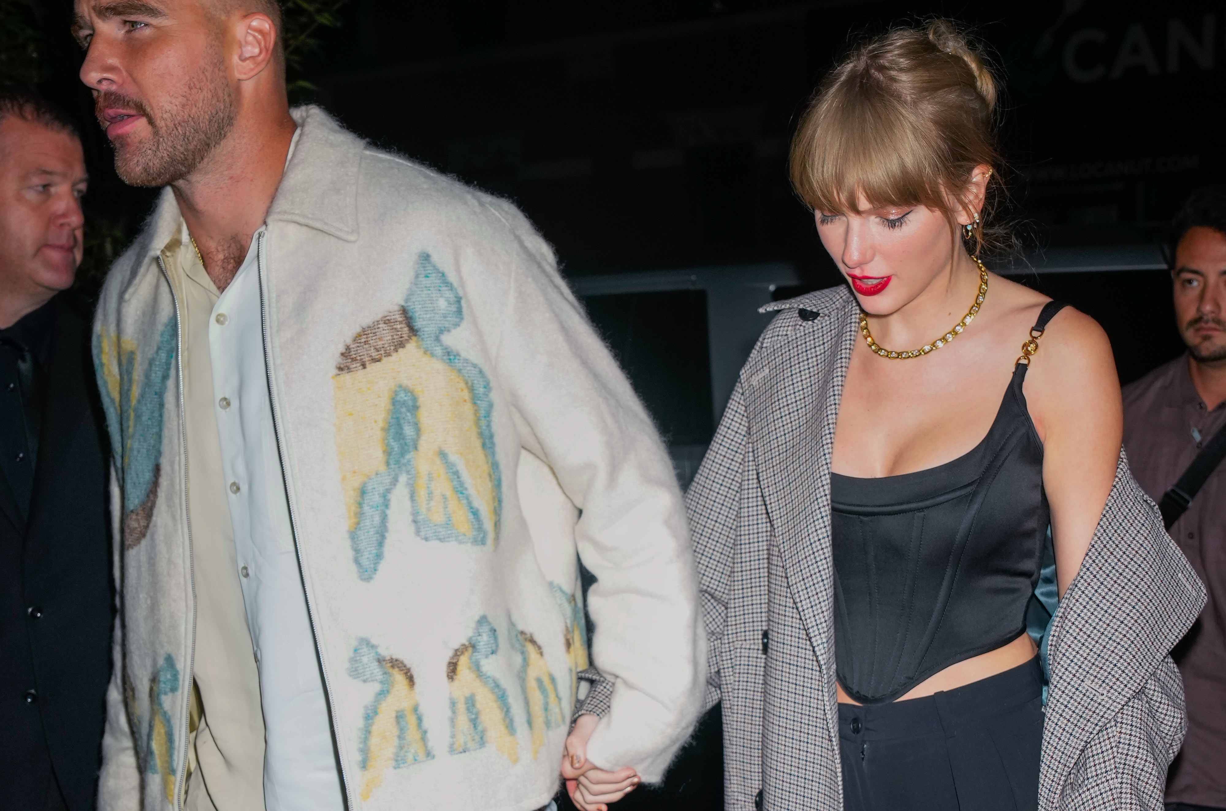 Closeup of Travis Kelce and Taylor Swift holding hands as they exit a building at night