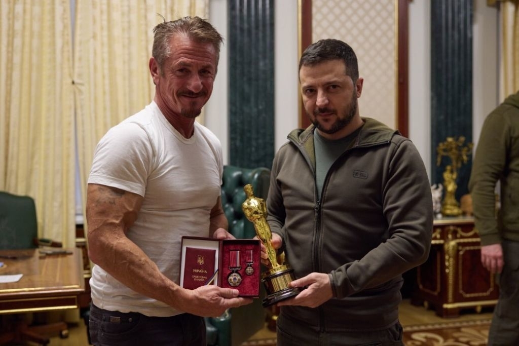 Sean Penn in a white t-shirt, standing with President Zelenskyy who is in a casual jacket, holding a trophy and award