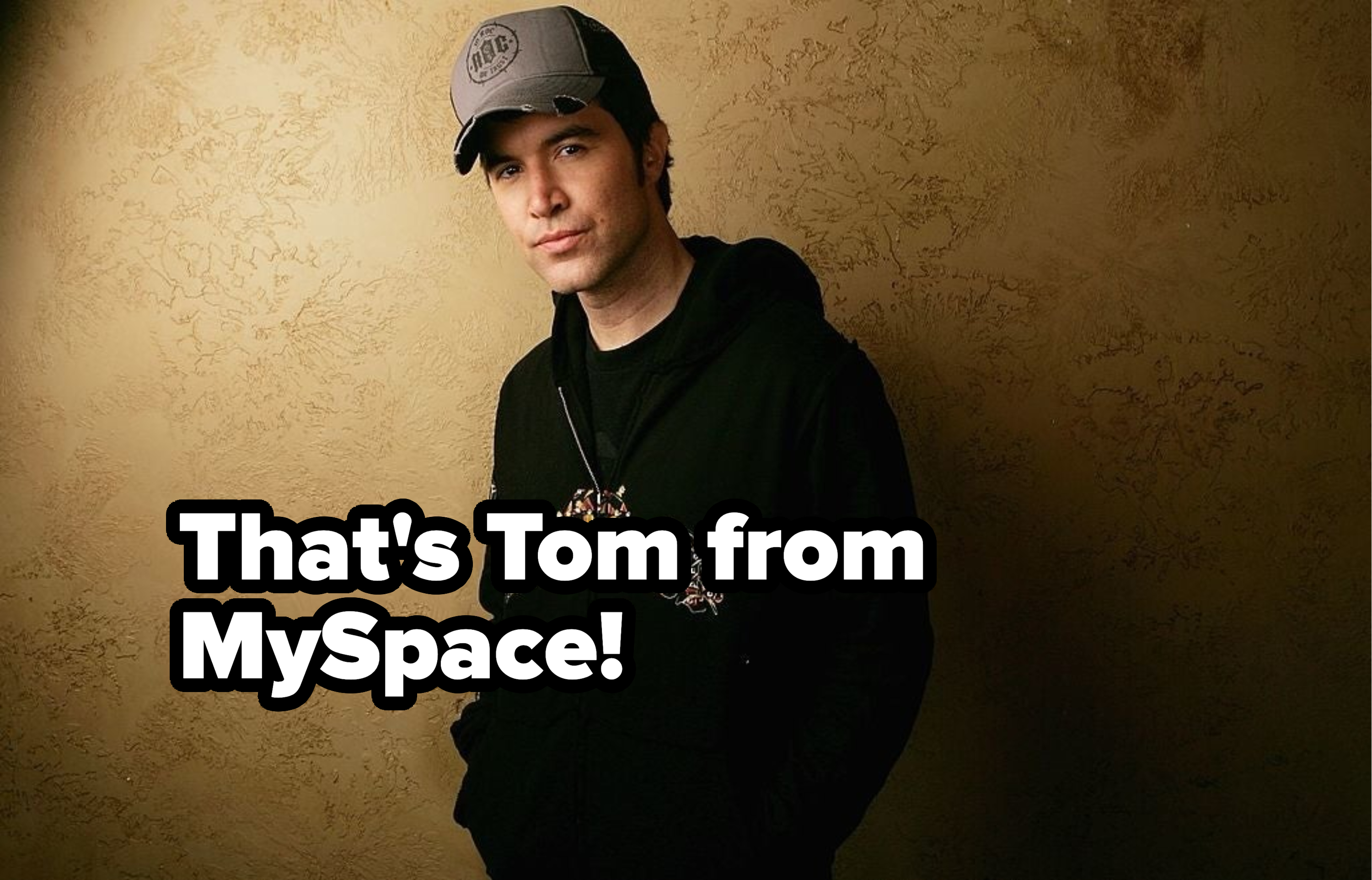 Man in cap and hoodie stands with hands in pockets against textured backdrop