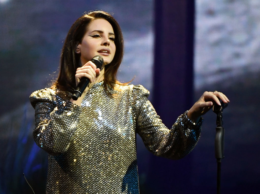Lana Del Rey performing on stage wearing a sparkling top, holding a microphone