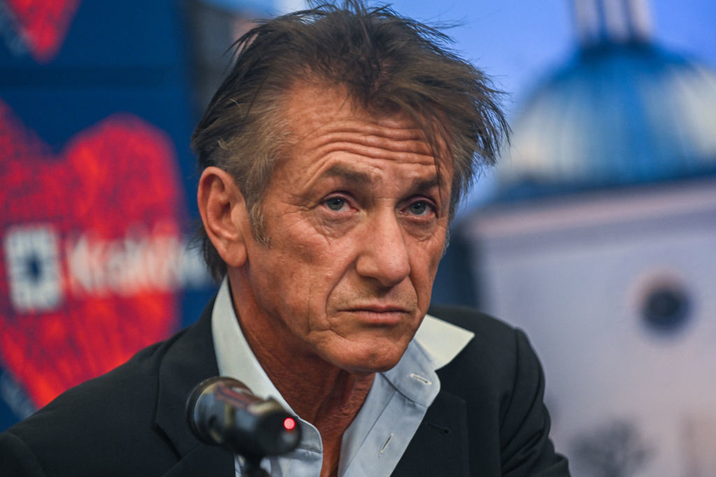 Sean Penn in a suit speaks at a microphone during an event