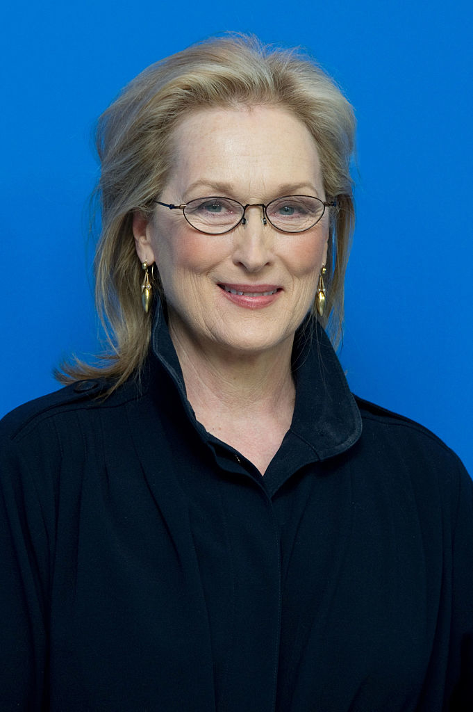 Meryl Streep smiling in a black outfit with glasses and earrings against a blue background