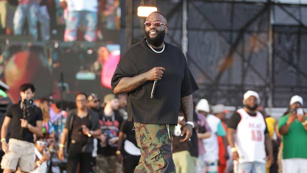 Rick Ross onstage performing, wearing a black shirt and camo pants with a backdrop of screens and audience