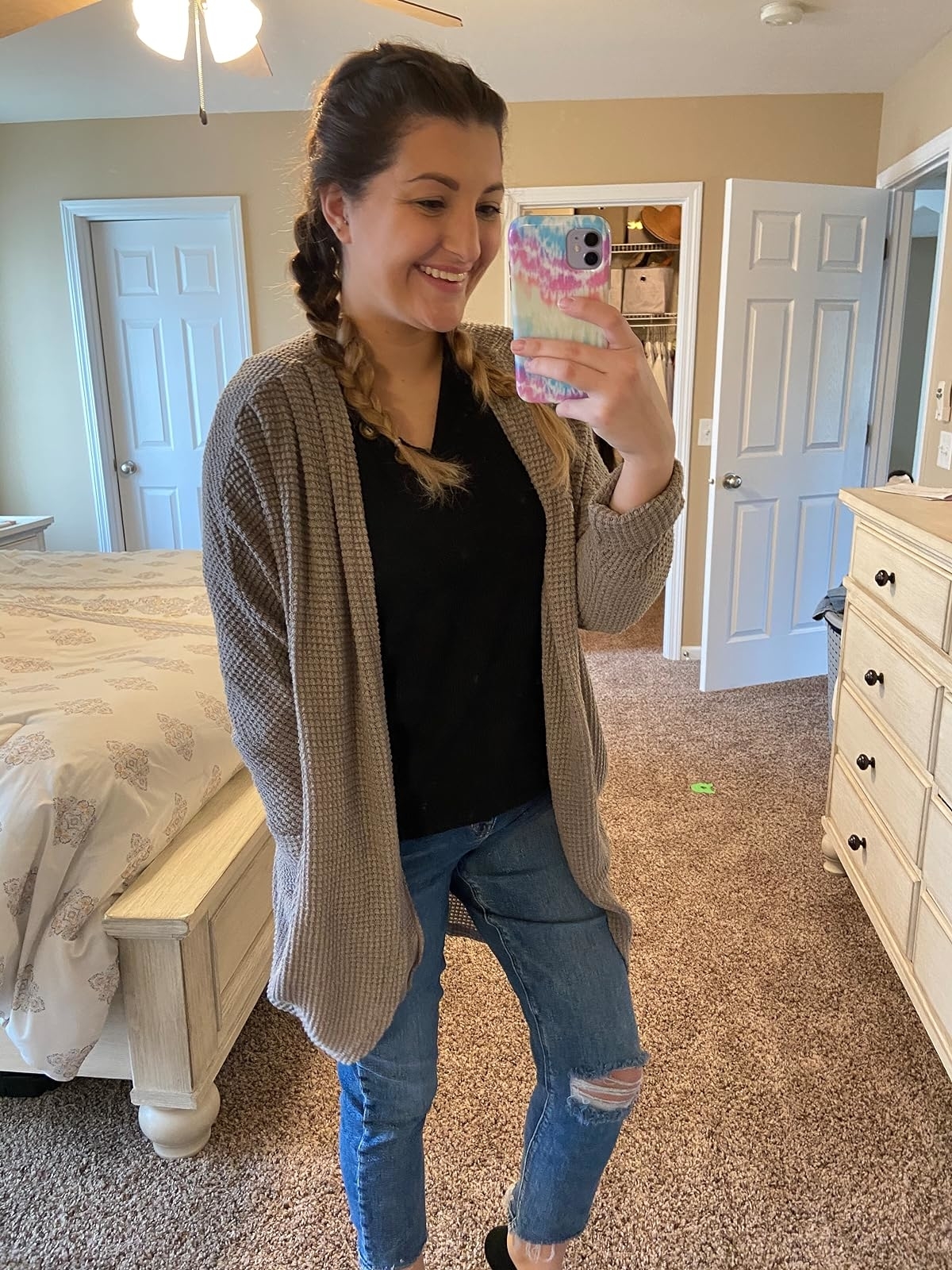 Reviewer's photo of them wearing the cardigan in the color grey, styled with jeans and a black t-shirt, taking a mirror selfie holding a phone with a patterned case