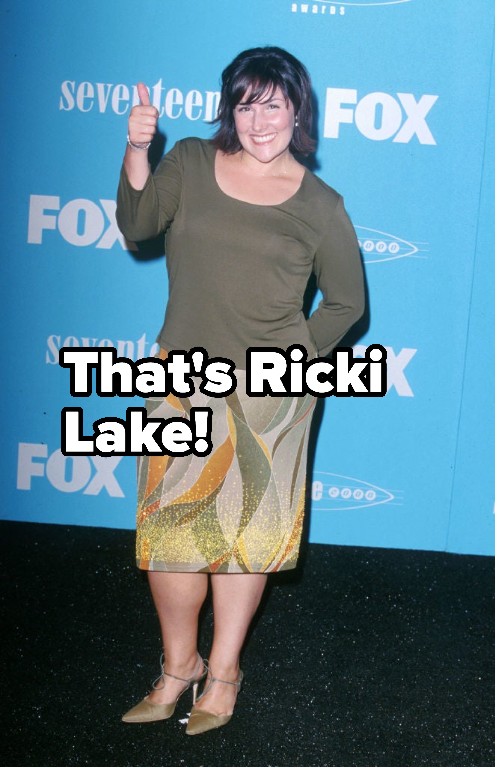 Woman poses with a thumbs-up, wearing a green top, patterned skirt, and beige shoes