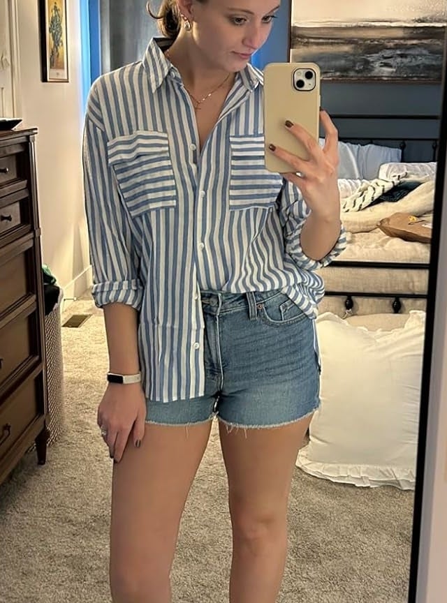 Reviewer&#x27;s photo of them wearing the button-down shirt in blue and white stripes, styled half-tucked into denim shorts, sleeves rolled to the elbows, and with white slide sandals, taking a mirror selfie
