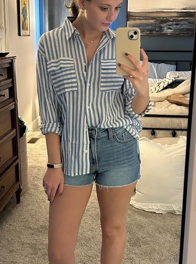 Reviewer's photo of them wearing the button-down shirt in blue and white stripes, styled half-tucked into denim shorts, sleeves rolled to the elbows, and with white slide sandals, taking a mirror selfie