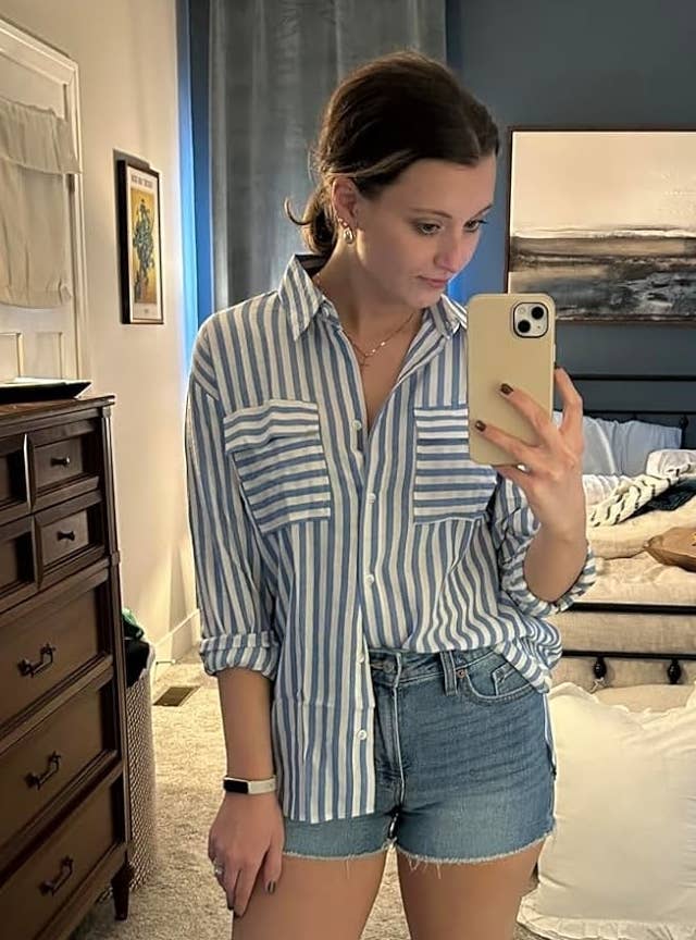 Reviewer's photograph of them wearing the button-down garment successful bluish and achromatic stripes, styled half-tucked into denim shorts, sleeves rolled to the elbows, and with achromatic descent sandals, taking a reflector selfie