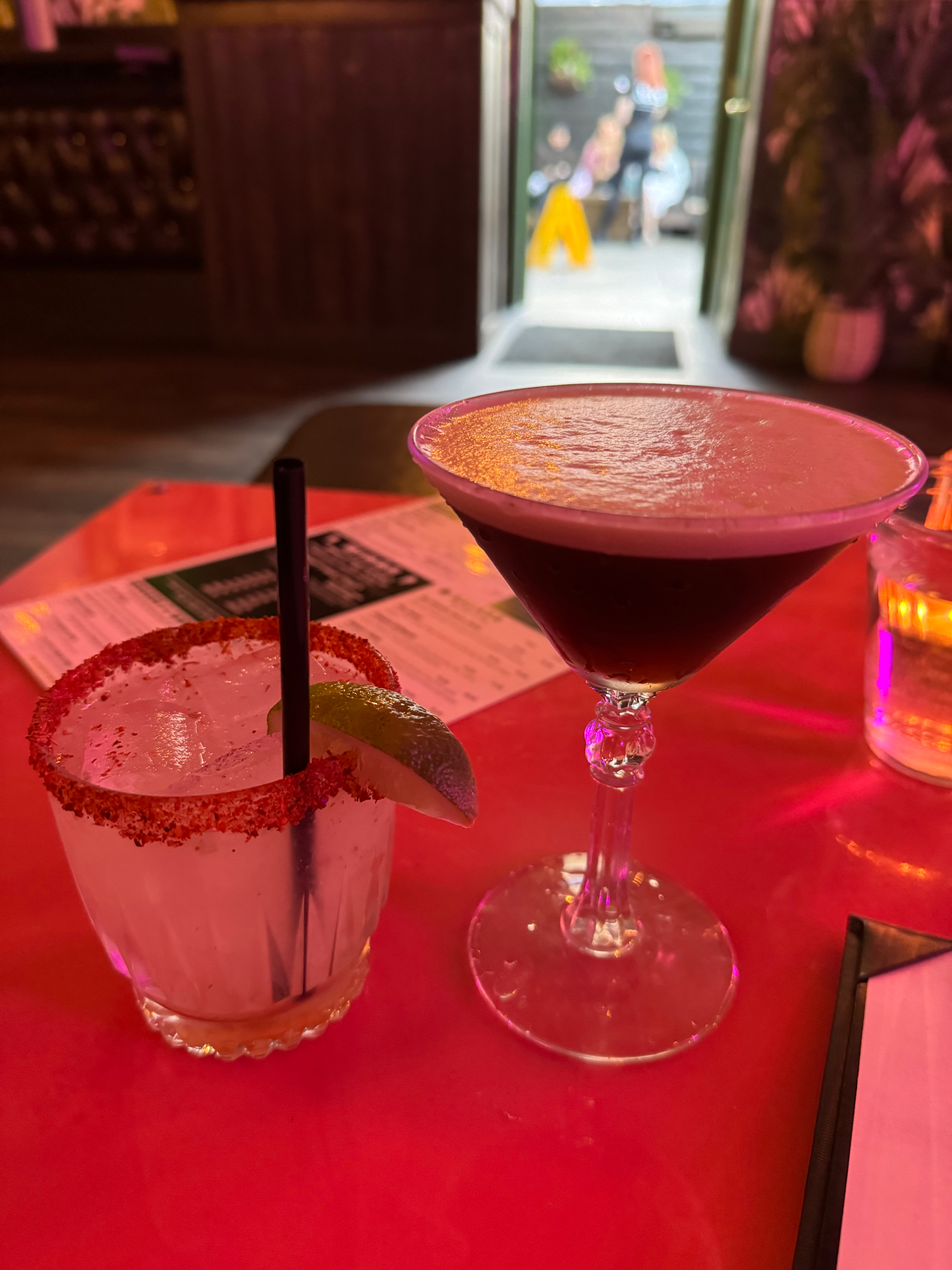 Two cocktails on a red table, one with a salted rim and lime wedge, the other a dark martini