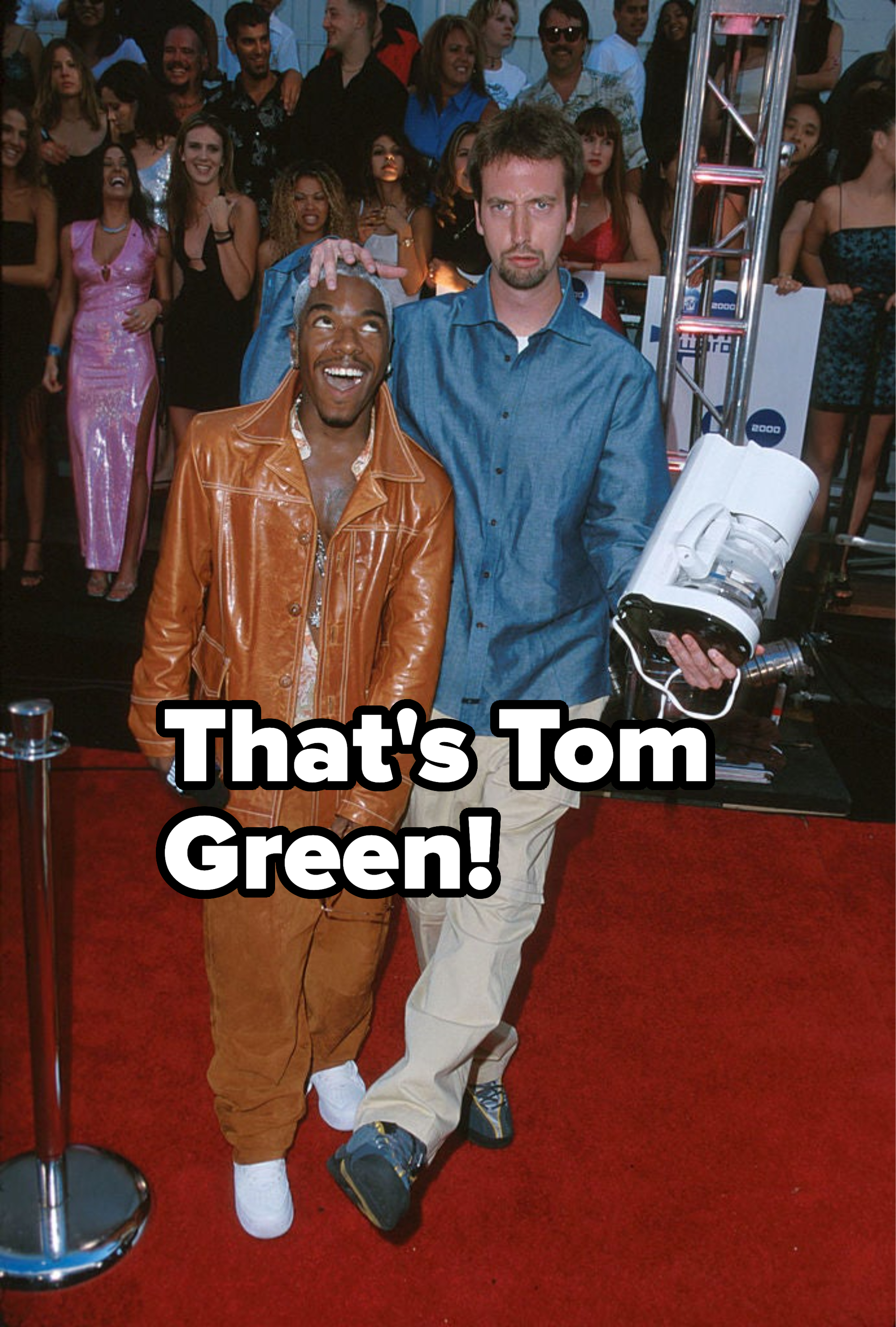 Two men on the red carpet, one in a leather jacket and pants, the other casual with a camera