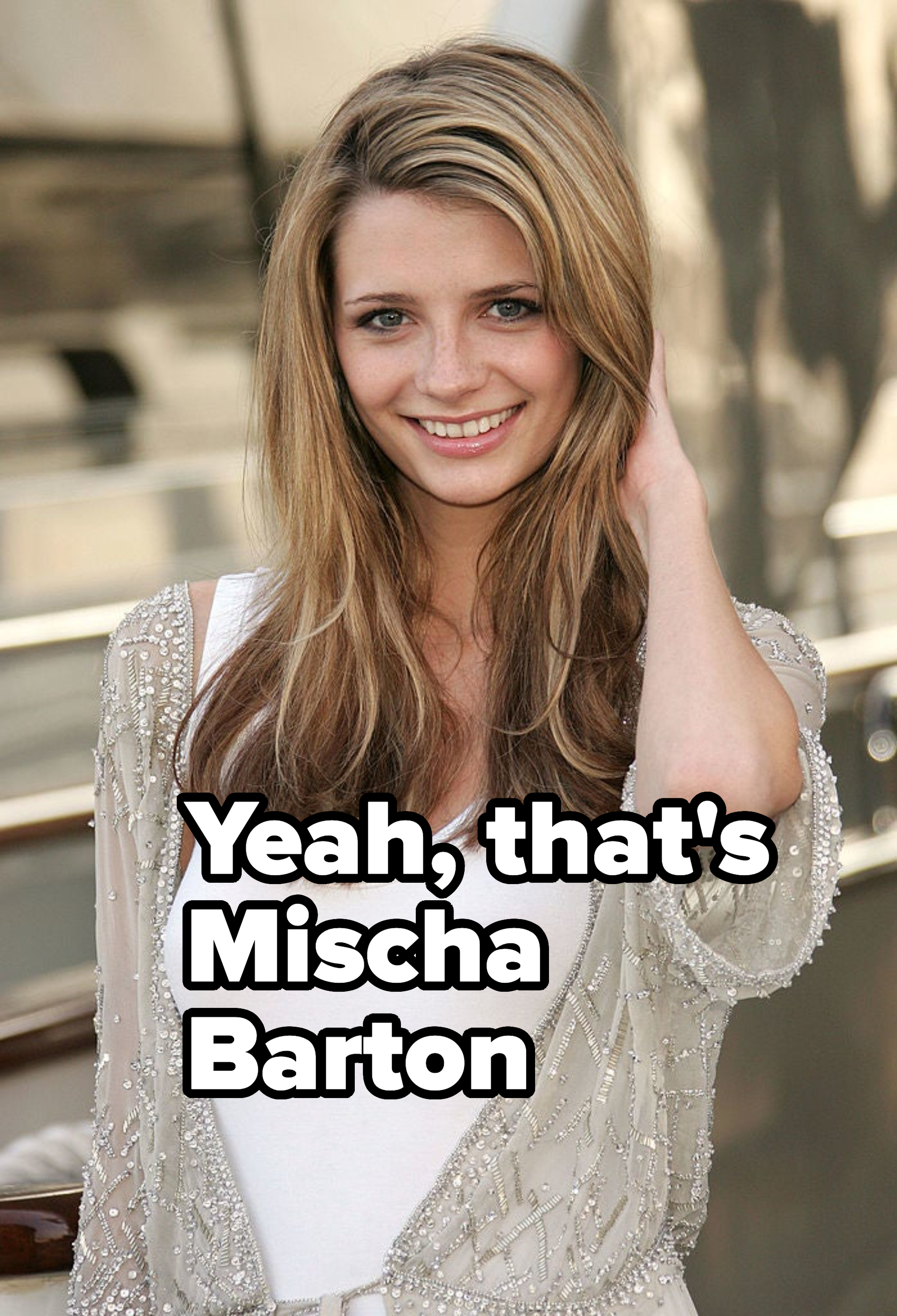 Actress smiling in a white top with beaded detailing, hand in hair, outdoors