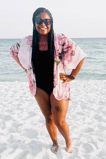 Reviewer's photograph of them wearing a pinkish floral coverup implicit a achromatic one-piece swimsuit connected a beauteous sandy formation with the water successful the background