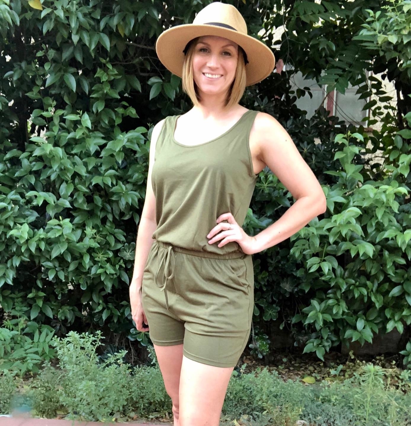 Reviewer&#x27;s photo of them wearing a sleeveless green romper, styled with a a straw sunhat and standing against a leafy green background
